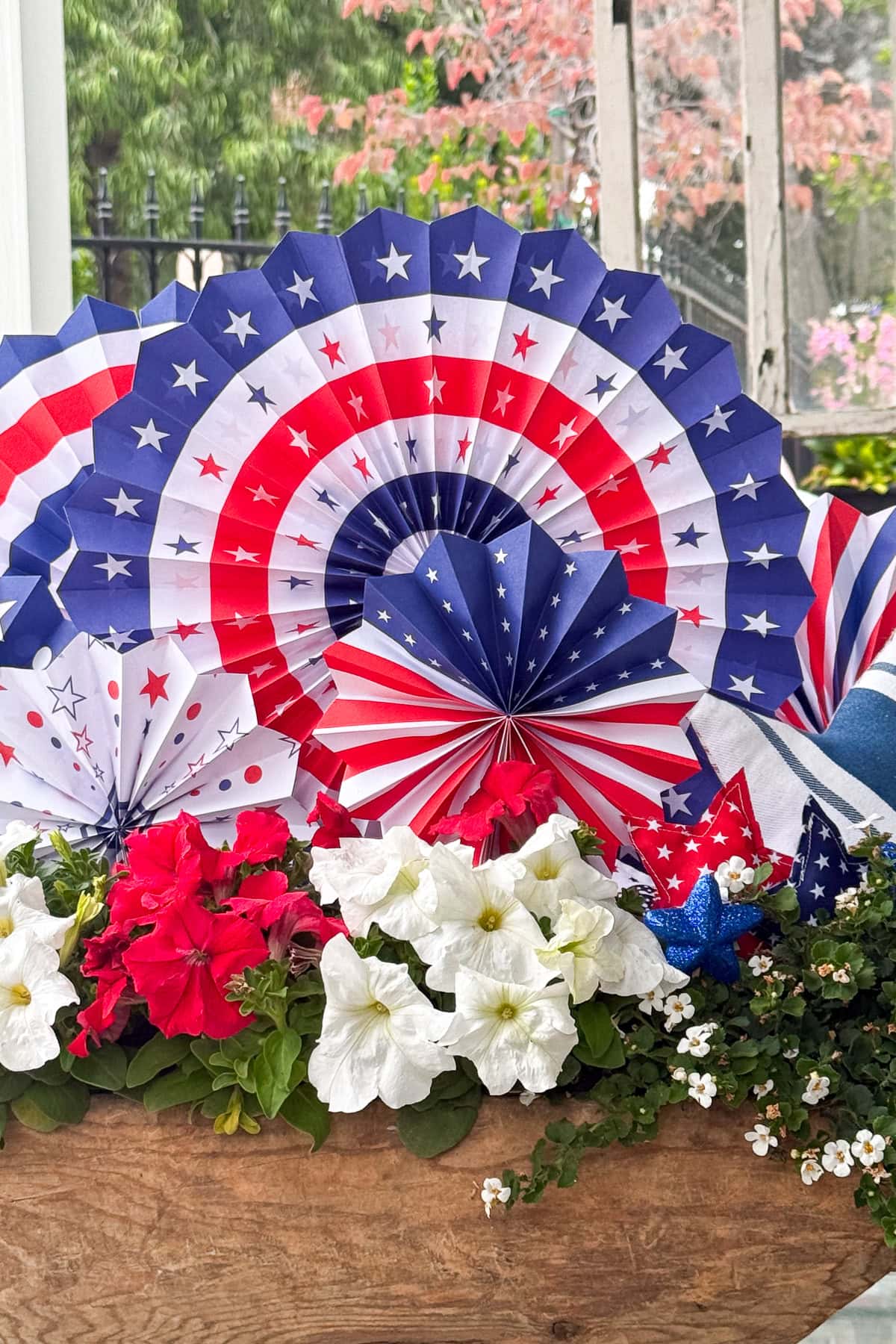 Red, white, and blue paper fans with stars and stripes decorate a planter filled with white and red flowers, creating a patriotic display. The background shows greenery and blurred pink foliage.