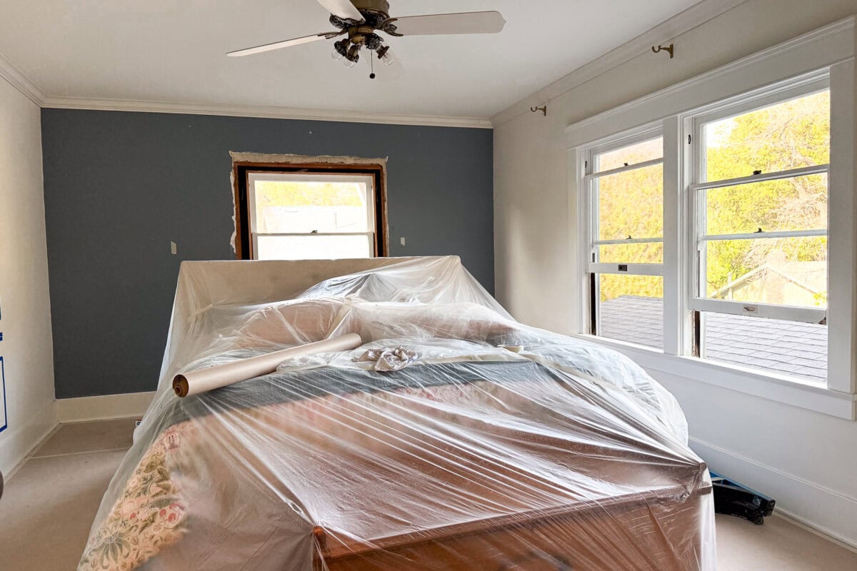 A bedroom under renovation with furniture covered in plastic sheeting. The wall is partially painted blue, and the window frame is unfinished. Sunlight streams through two large windows on the right.