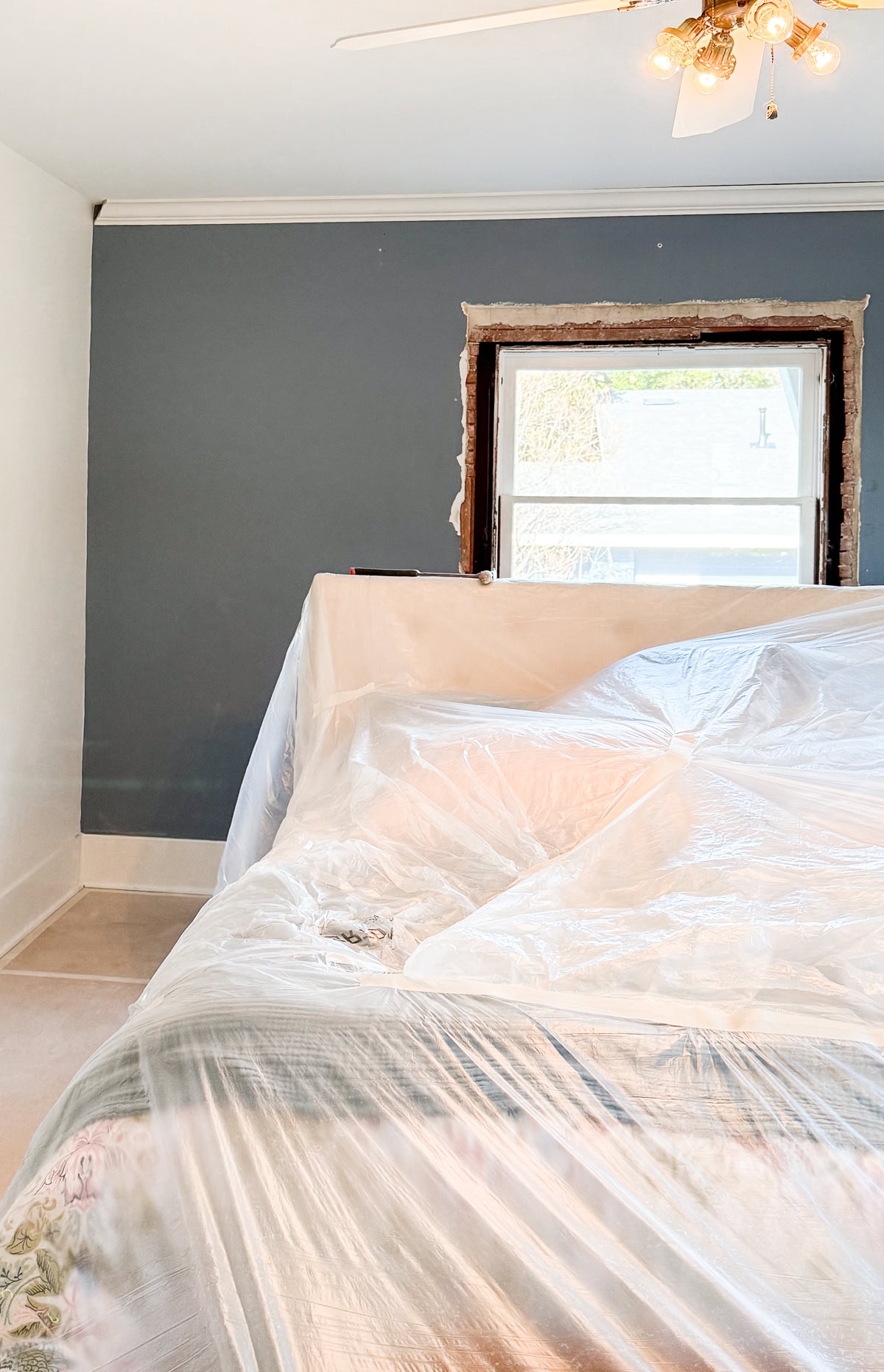 A bedroom before a renovation with a bed covered in clear plastic sheeting, likely for protection during renovation. The window frame is unfinished, and the walls are painted blue and white. A ceiling fan with lights is visible.