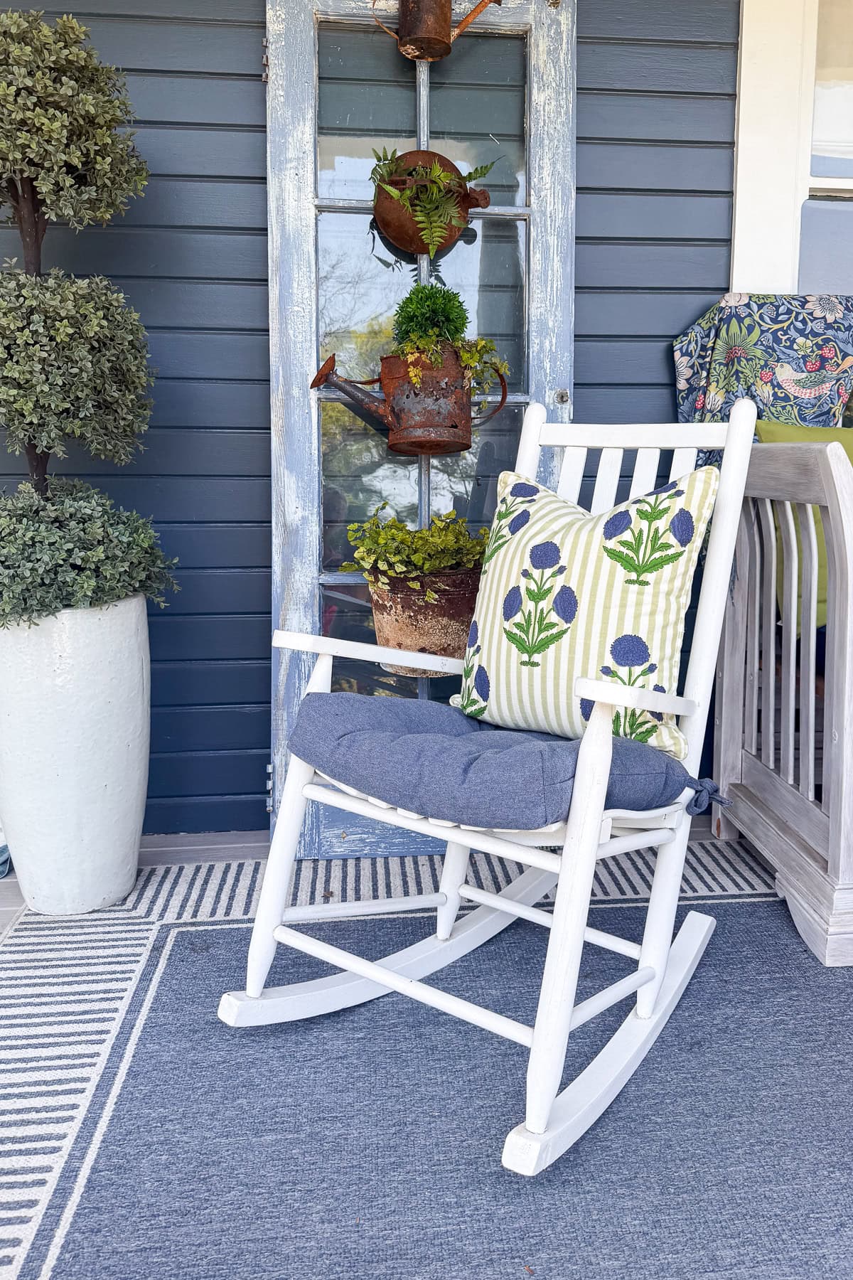 A white rocking chair with a blue cushion and floral pillow sits on a blue and white rug in front of a vintage door. A large potted plant and vertical planter add easy porch decor, perfect for refreshing your summer porch decorations.