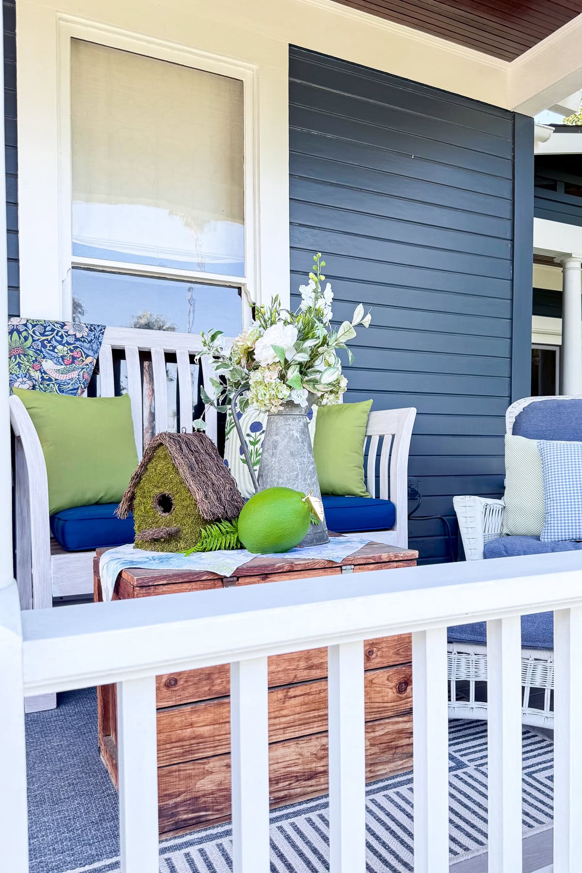 A cozy porch with a white bench, blue and green cushions, a wooden chest as a table, a birdhouse, a green ball, and a vase of white flowers. The house has blue siding and white trim.