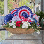 A wooden planter holds white and red flowers with greenery, decorated with red, white, and blue paper fans and small American flags, set on a glass table outdoors.