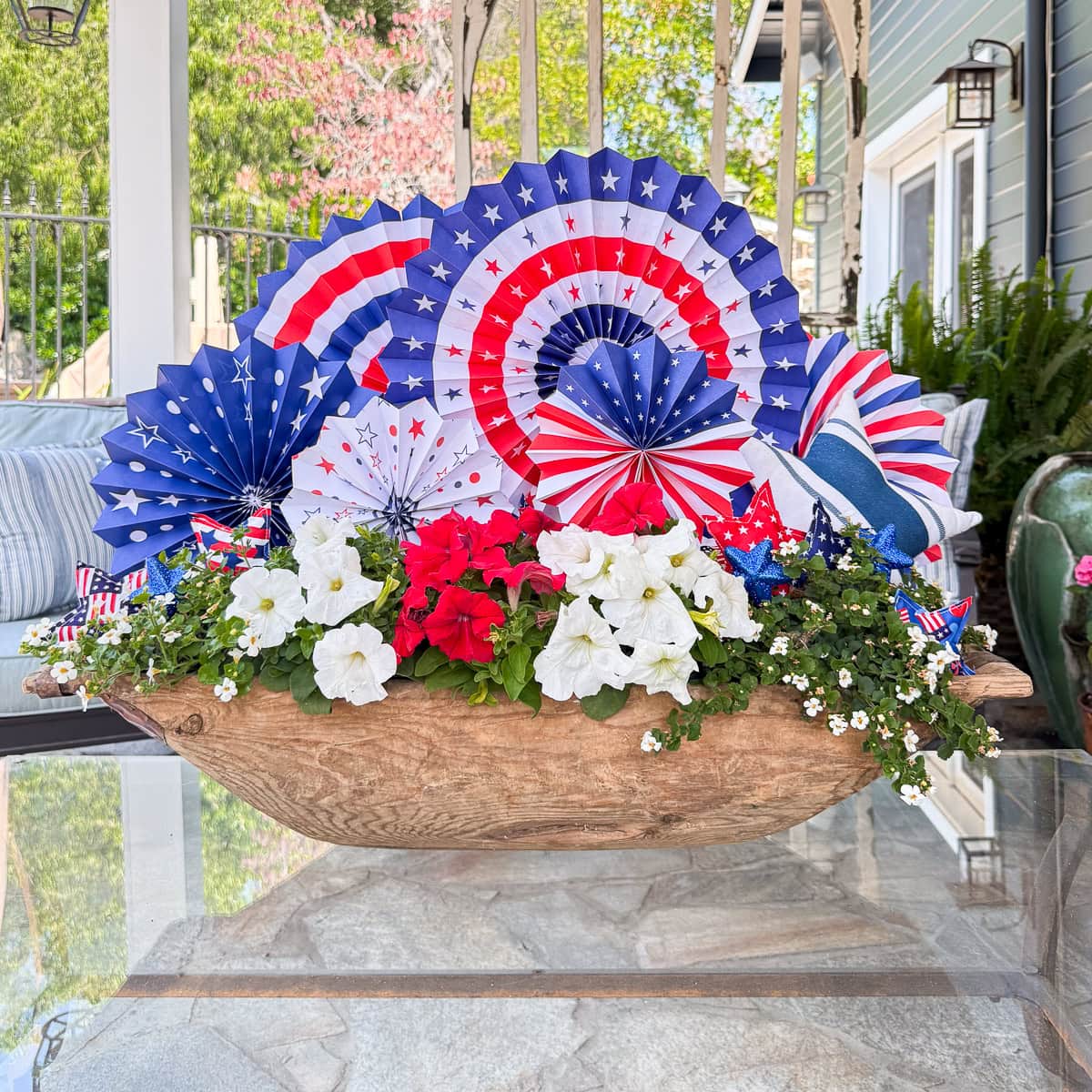 A wooden planter holds white and red flowers, green foliage, and is decorated with festive red, white, and blue paper fans and stars, creating a patriotic display on an outdoor glass table.