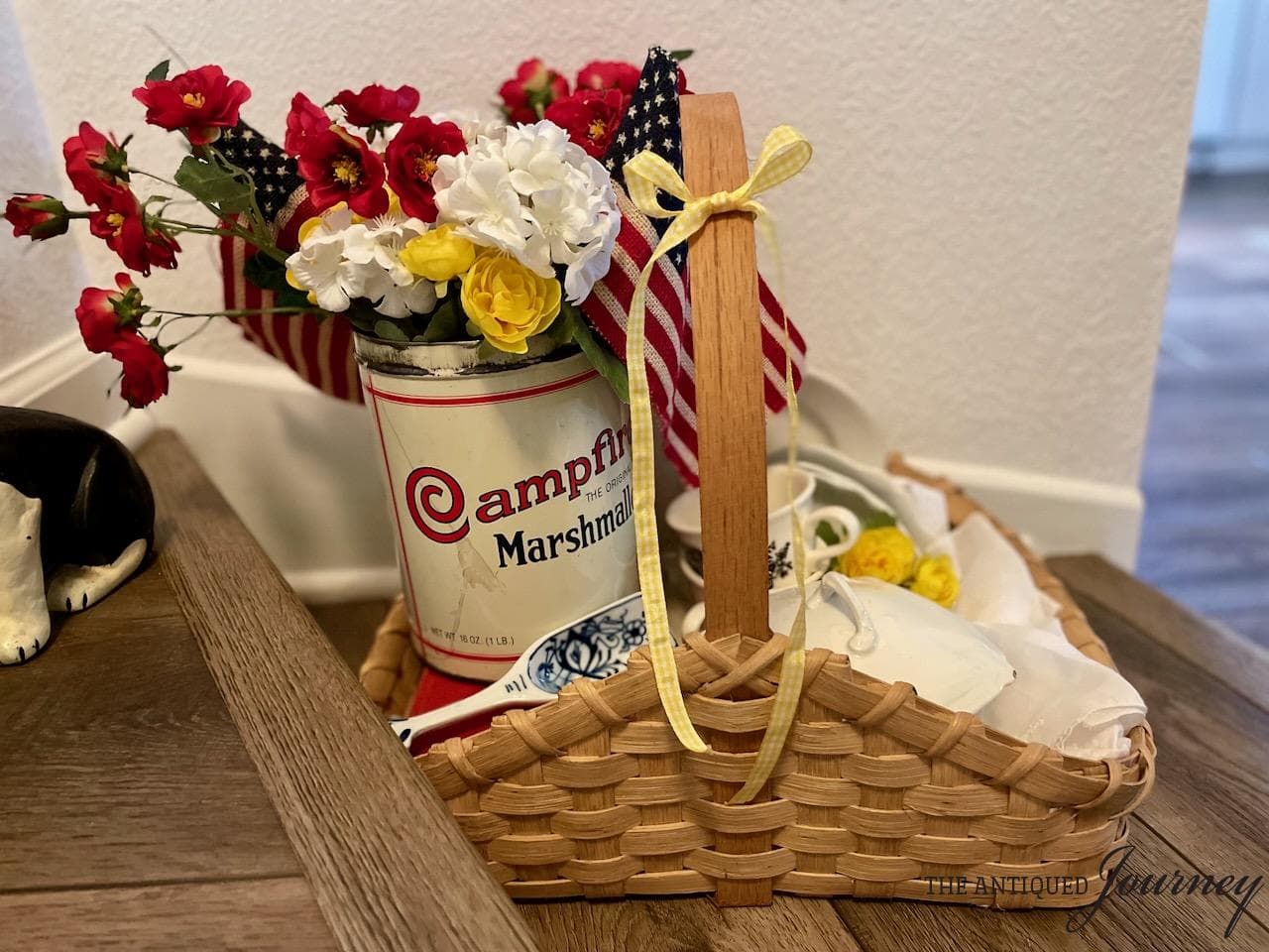 A charming basket centerpiece features a tin can with Campfire Marshmallows branding, filled with red, white, and yellow flowers, small American flags, and teacups on a wooden floor near a white wall—perfect for the 4th of July.