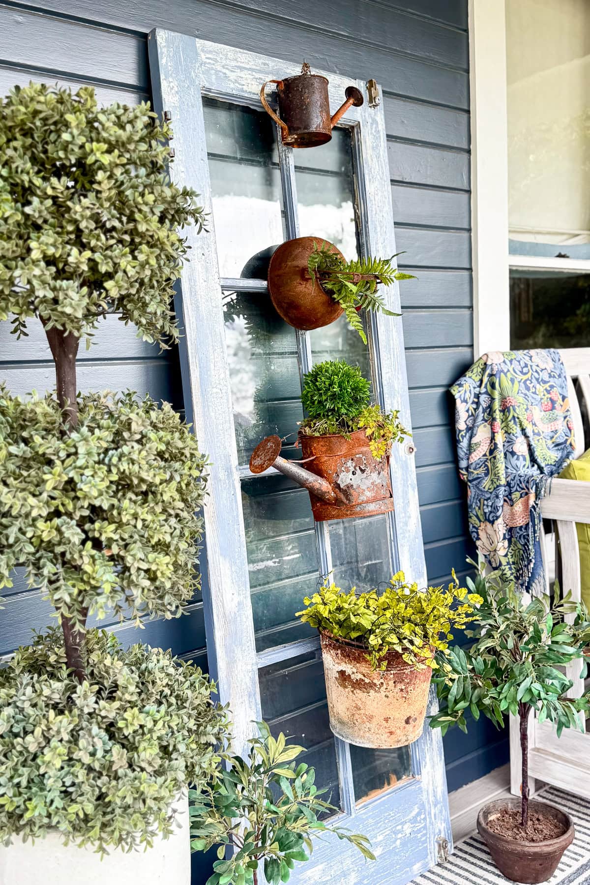 A rustic blue door with glass panes leans against a blue wall, decorated with potted plants and vintage watering cans. Nearby are potted topiary trees and a patterned cloth draped over a porch chair.