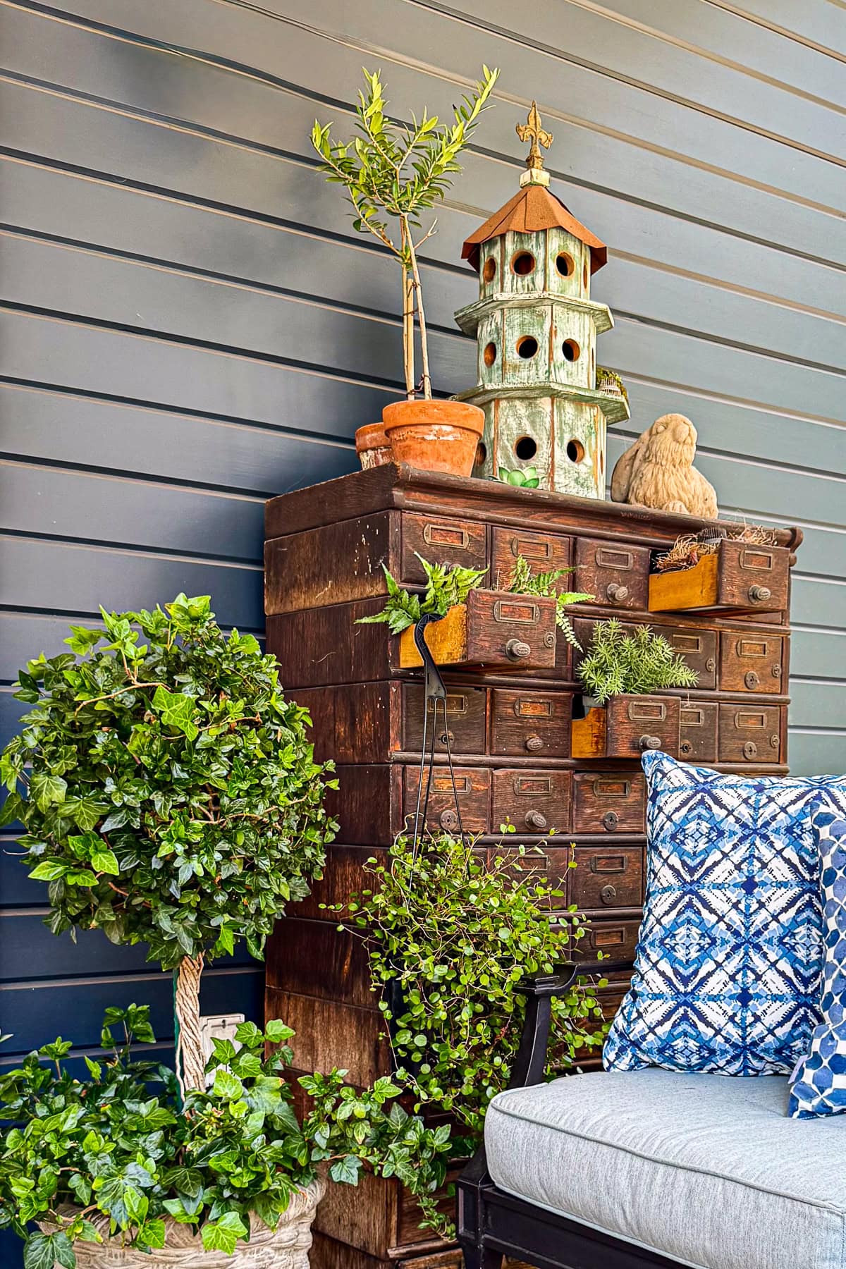 A vintage wooden apothecary cabinet with open drawers holds potted plants and greenery. A decorative birdhouse and sculpture sit on top. Beside it is a leafy potted plant and a bench with blue-patterned pillows.