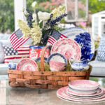 A wicker basket holds vintage red, white, and blue dishes, a floral arrangement, small American flags, and Cracker Jack boxes. More dishware and patriotic pillows decorate the background, creating a festive, summery display.