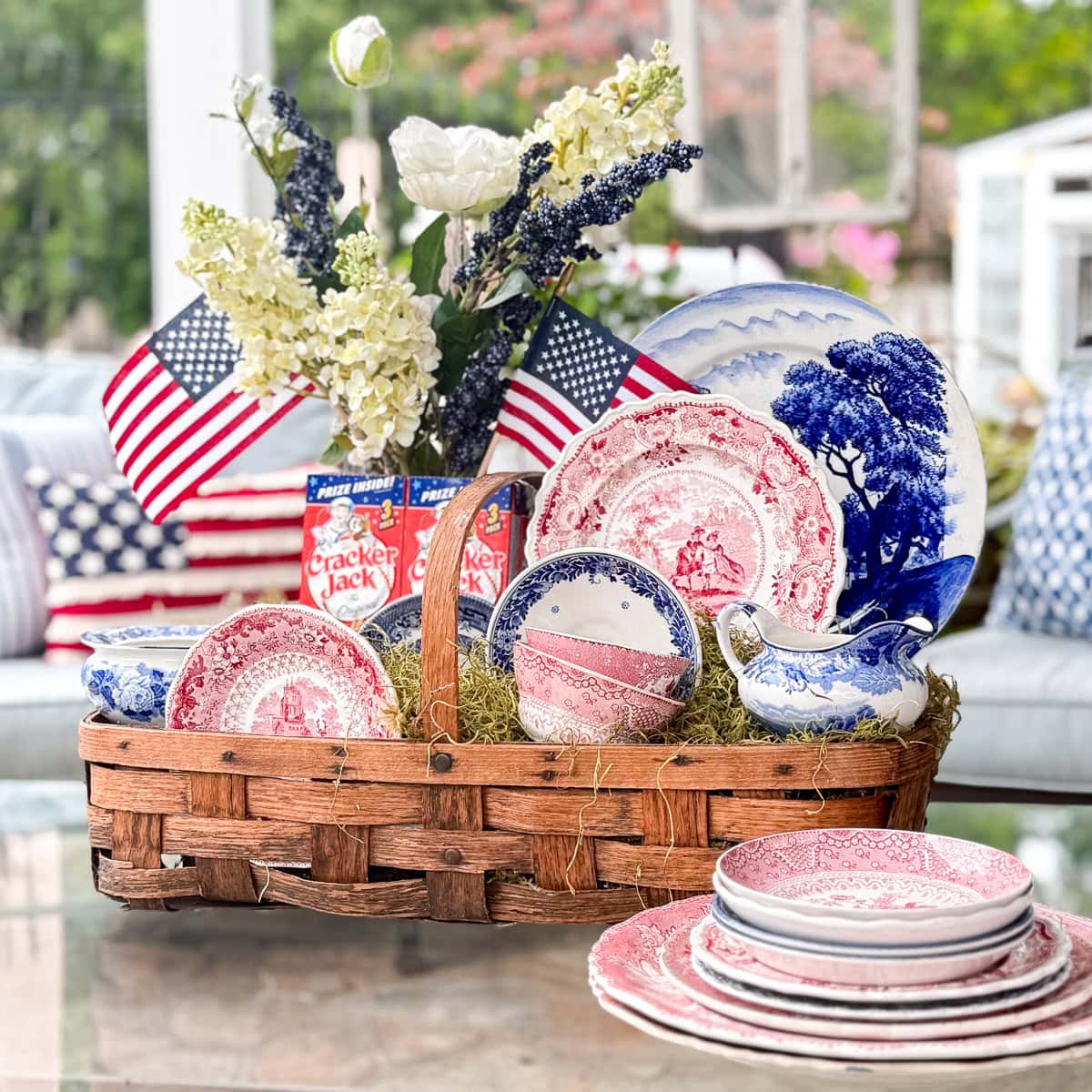 A wicker basket holds vintage red, white, and blue dishes, a floral arrangement, small American flags, and Cracker Jack boxes. More dishware and patriotic pillows decorate the background, creating a festive, summery display.