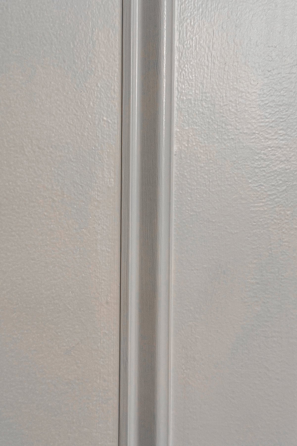Close-up of a gray textured wall with a vertical strip of trim or molding in the center, separating two slightly different shades of gray. The surface has a subtle shine and visible brushstroke patterns.
