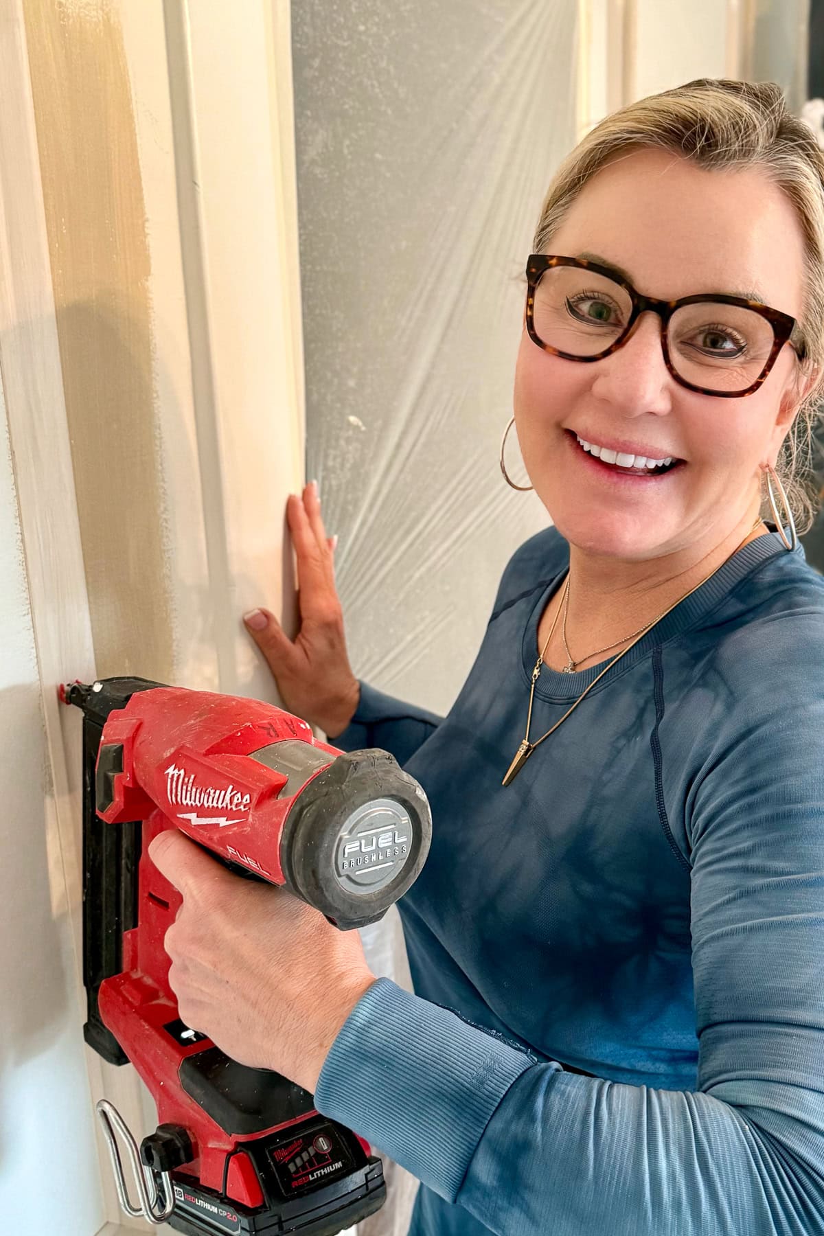 A smiling woman wearing glasses and a blue shirt uses a red Milwaukee power tool to work on a wall, with protective plastic covering in the background.