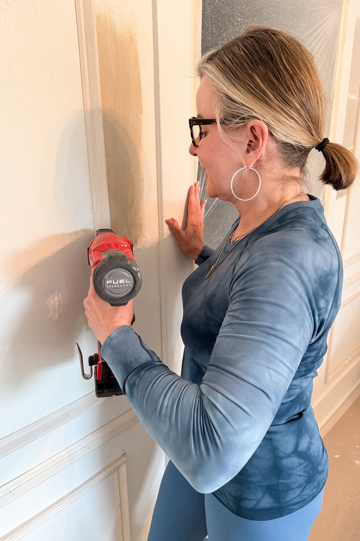 A woman wearing glasses and hoop earrings uses a red power sander on a white wall, smoothing the surface. She is dressed in a blue long-sleeve shirt and has her hair tied back in a ponytail.