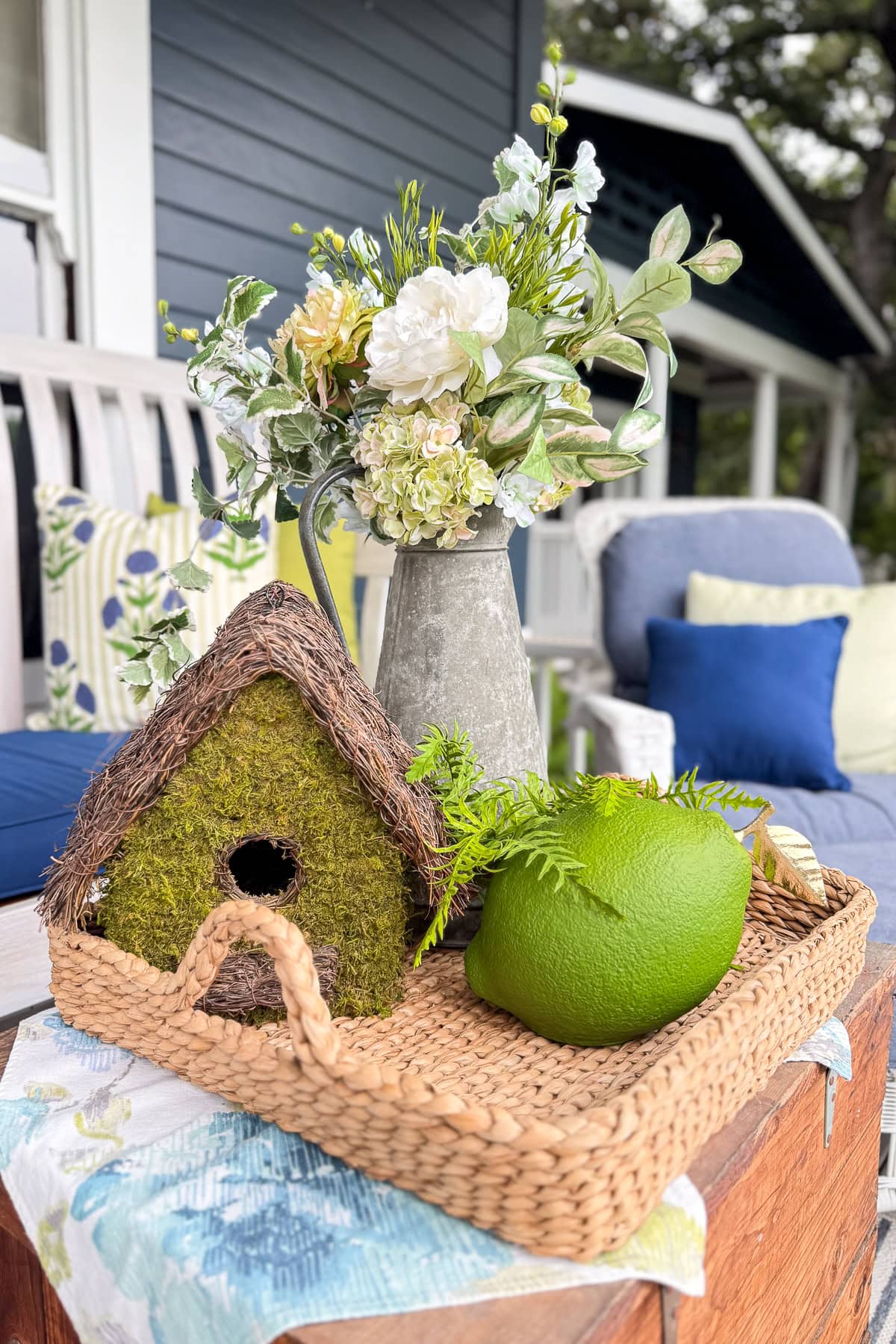 A wicker tray holds a mossy birdhouse, a green fruit, and a metal pitcher with white flowers on an outdoor table. Cushioned patio seating and a blue house exterior are visible in the background.