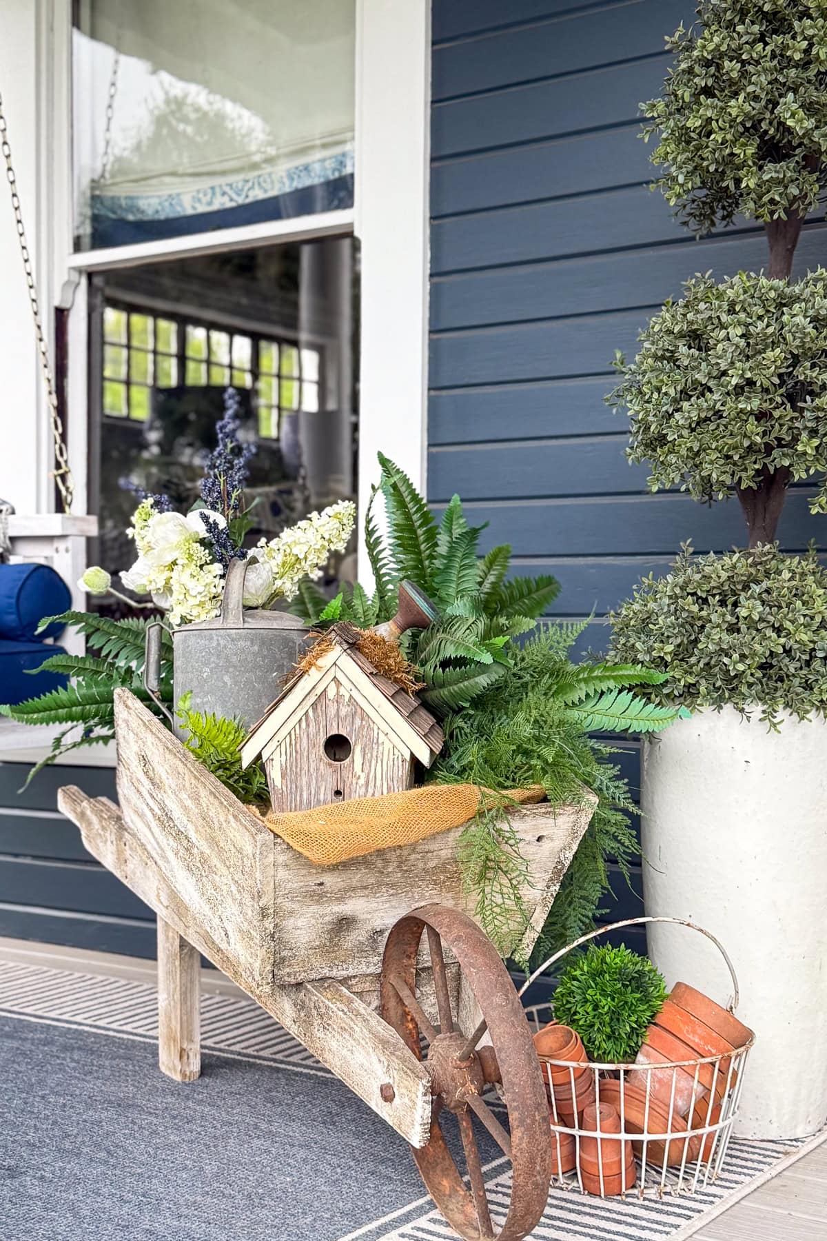 A rustic wooden wheelbarrow filled with green ferns, a birdhouse, a metal watering can, and a small mat sits on a porch beside a potted topiary and a white basket holding small clay flowerpots.