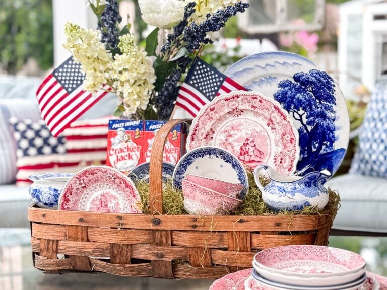 A 4th of July basket centerpiece features a wicker basket filled with red, white, and blue vintage dishes on a table, surrounded by American flags, Cracker Jack boxes, and a bouquet of white flowers in the background.