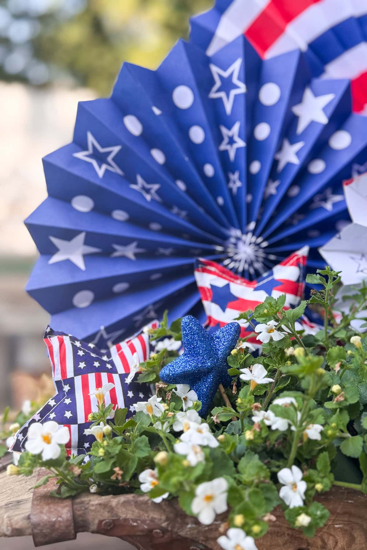 Patriotic decorations with blue and red paper fans featuring stars and stripes, a blue glittery star, and white flowers in the foreground.