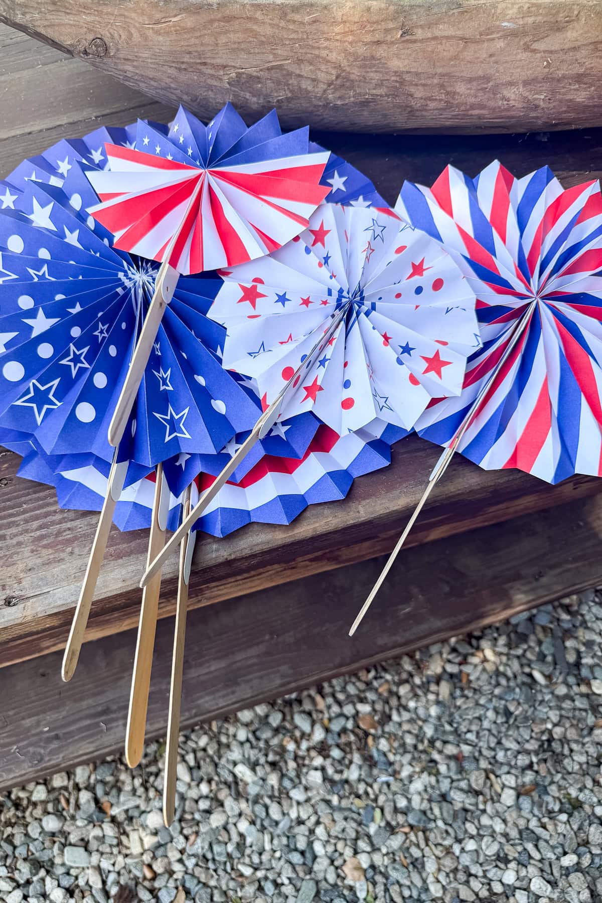 Paper fans in red, white, and blue designs with stars, stripes, and polka dots are displayed on wooden steps, with gravel beneath. The fans are attached to wooden sticks.