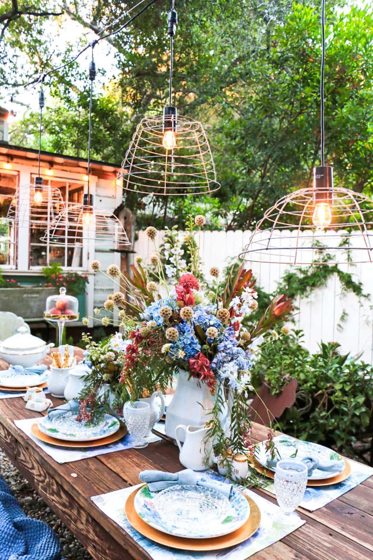 A rustic outdoor dining table set with blue and white plates, glassware, and a large floral centerpiece, beneath hanging cage pendant lights, surrounded by greenery and a white fence.