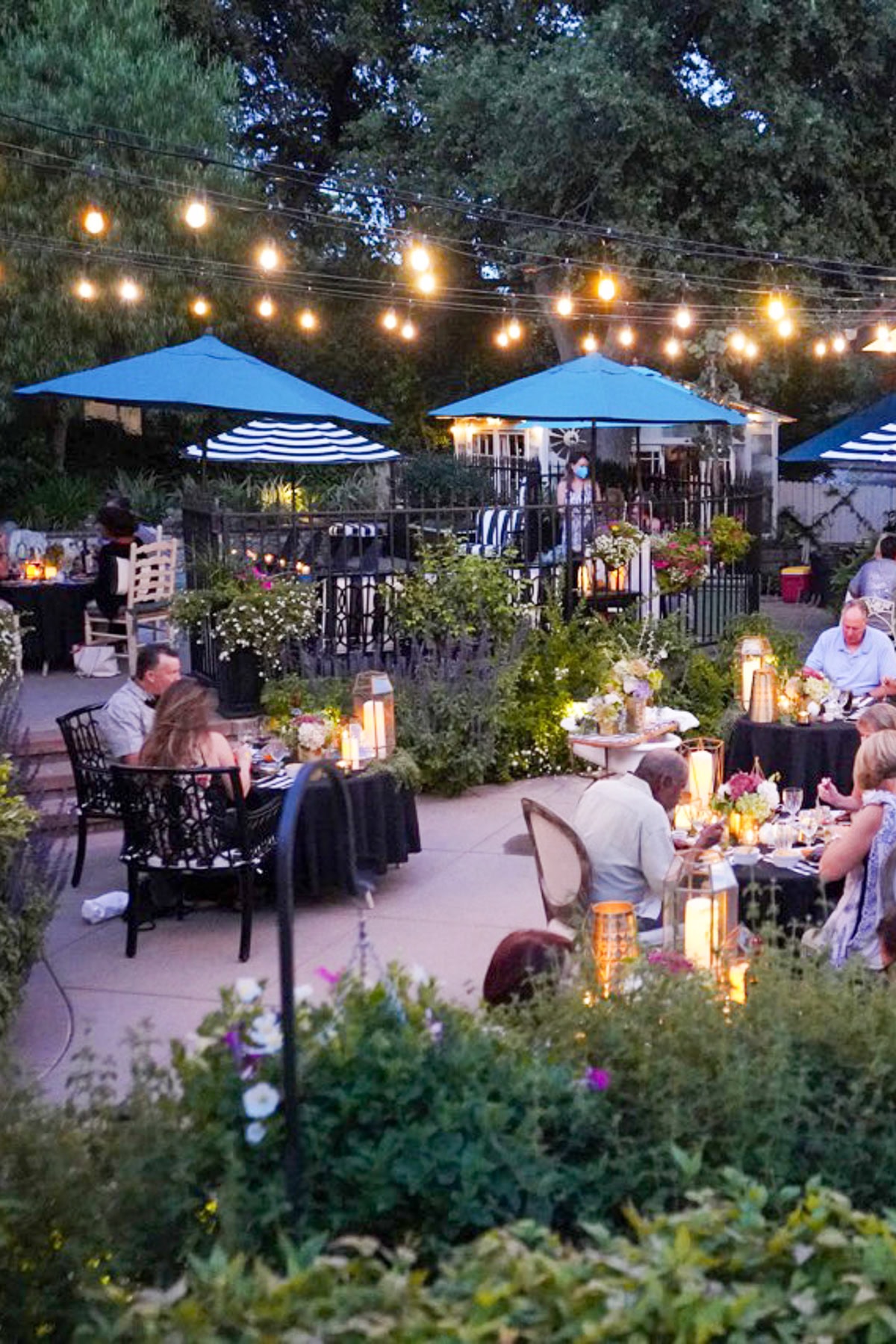 People are dining outdoors at night under string lights and blue umbrellas, surrounded by lush greenery and blooming flowers. Tables are set with food, drinks, and lanterns, creating a cozy, festive atmosphere.