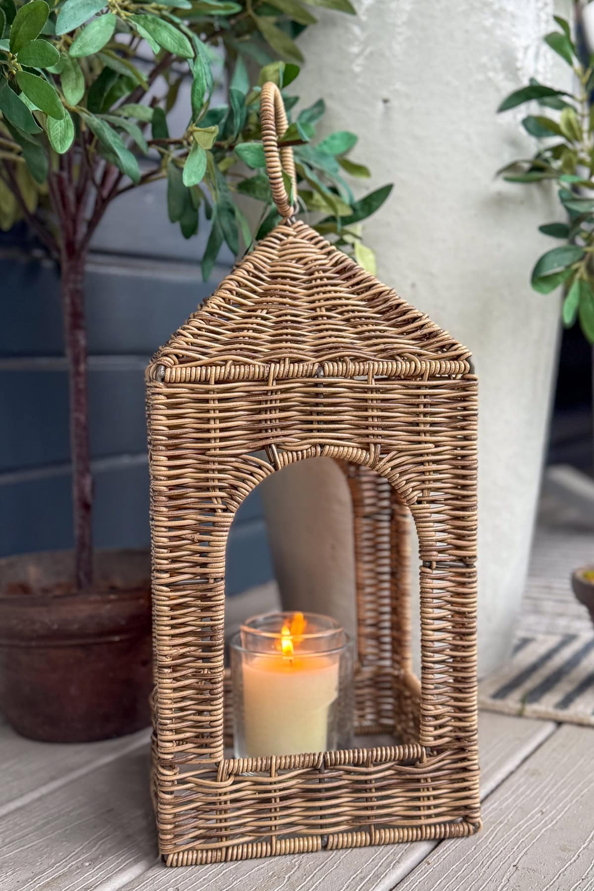 A candle in a glass holder sits inside a house-shaped wicker lantern on a wooden surface, with potted plants and a large white vase in the background.