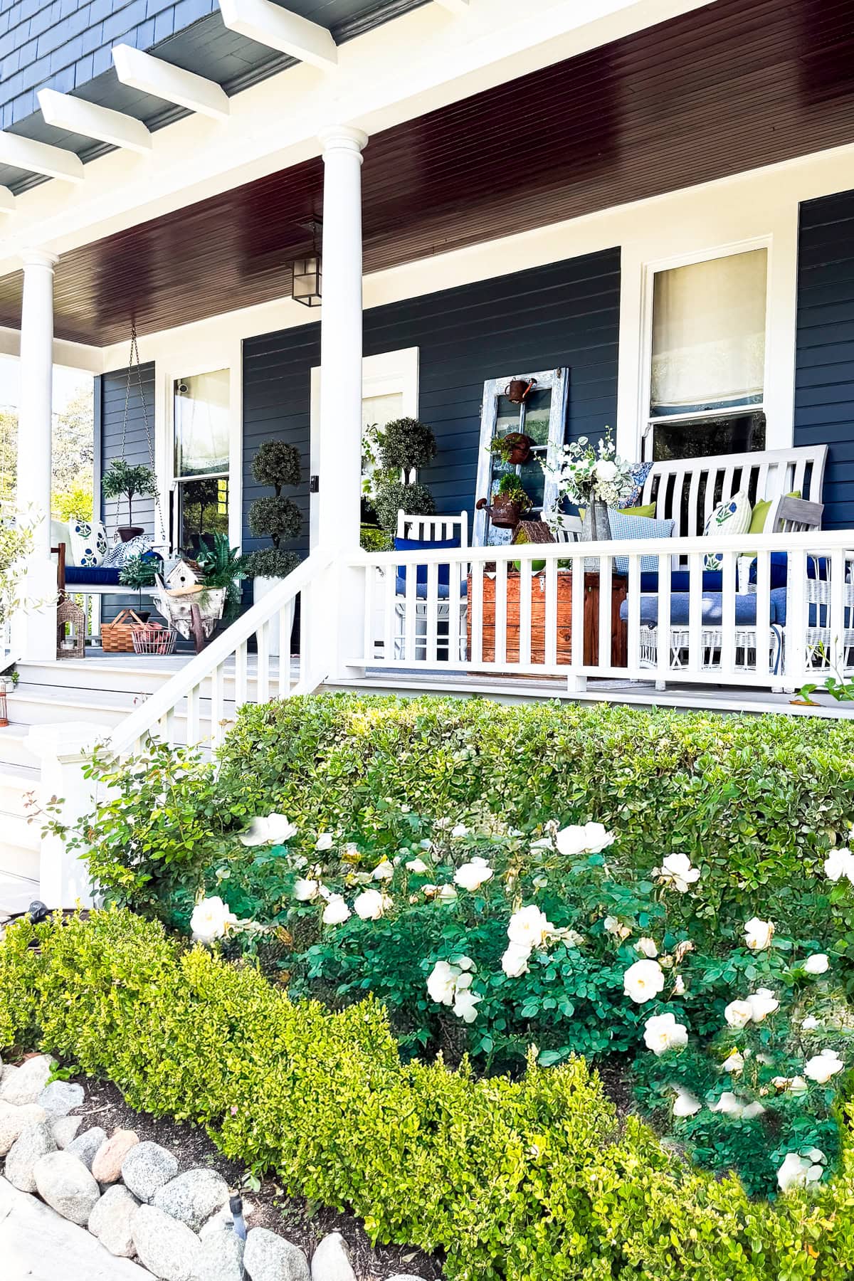 A charming front porch with white railings, rocking chairs, and potted plants features easy summer porch decor—blooming white roses and green hedges bordered by a stone edge offer refreshing porch decorating ideas.