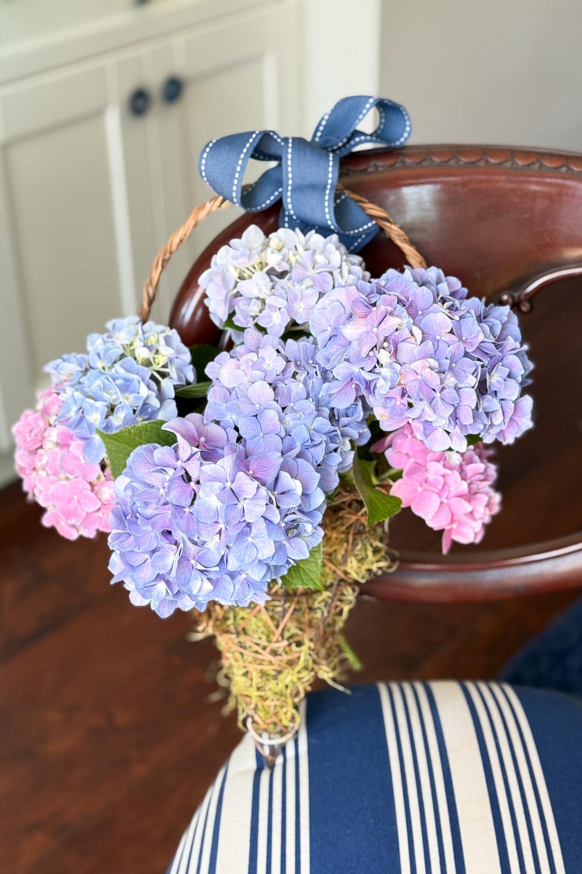 A cone-shaped wicker basket filled with purple, blue, and pink hydrangeas, topped with a blue ribbon, hangs on the back of a wooden chair with striped upholstery.