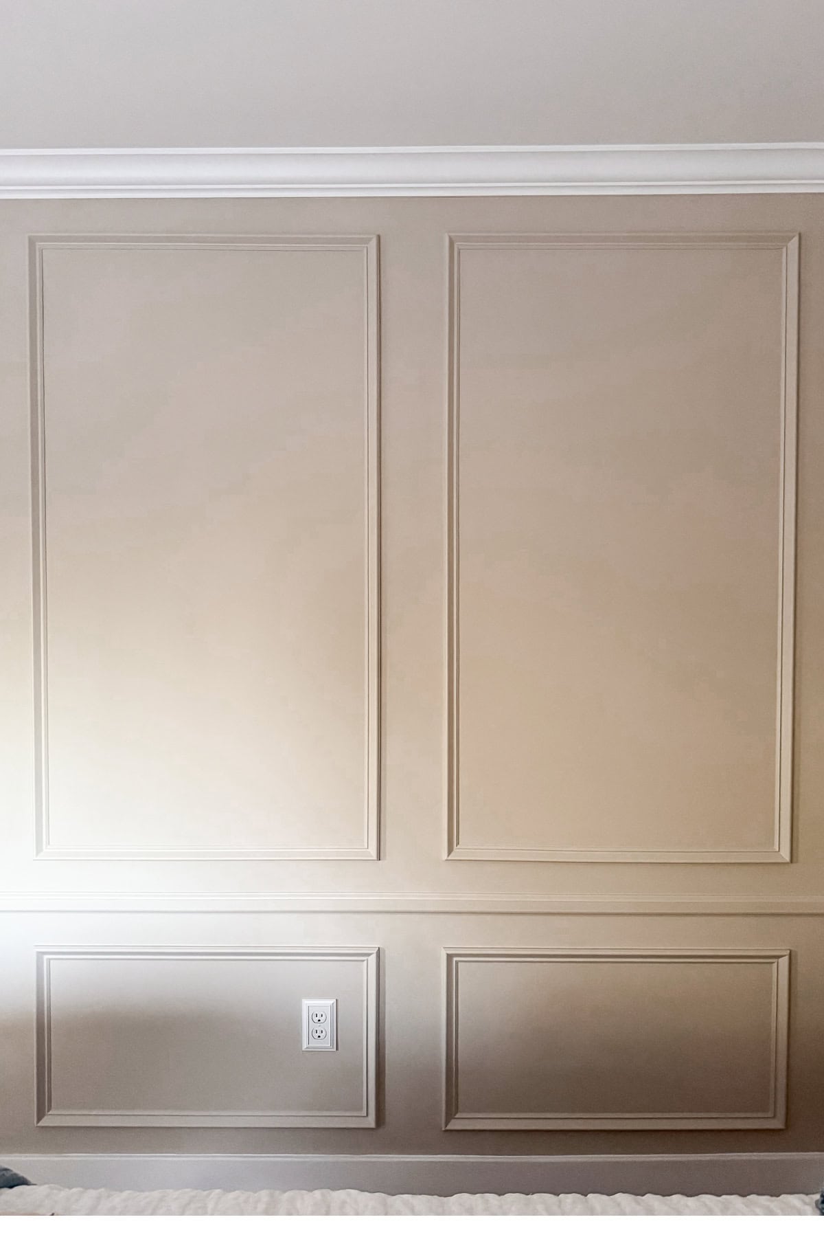 A beige wall with DIY wall moldings featuring decorative rectangular panels, white crown molding at the top, an electrical outlet near the bottom, and part of a white textured surface along the bottom edge.