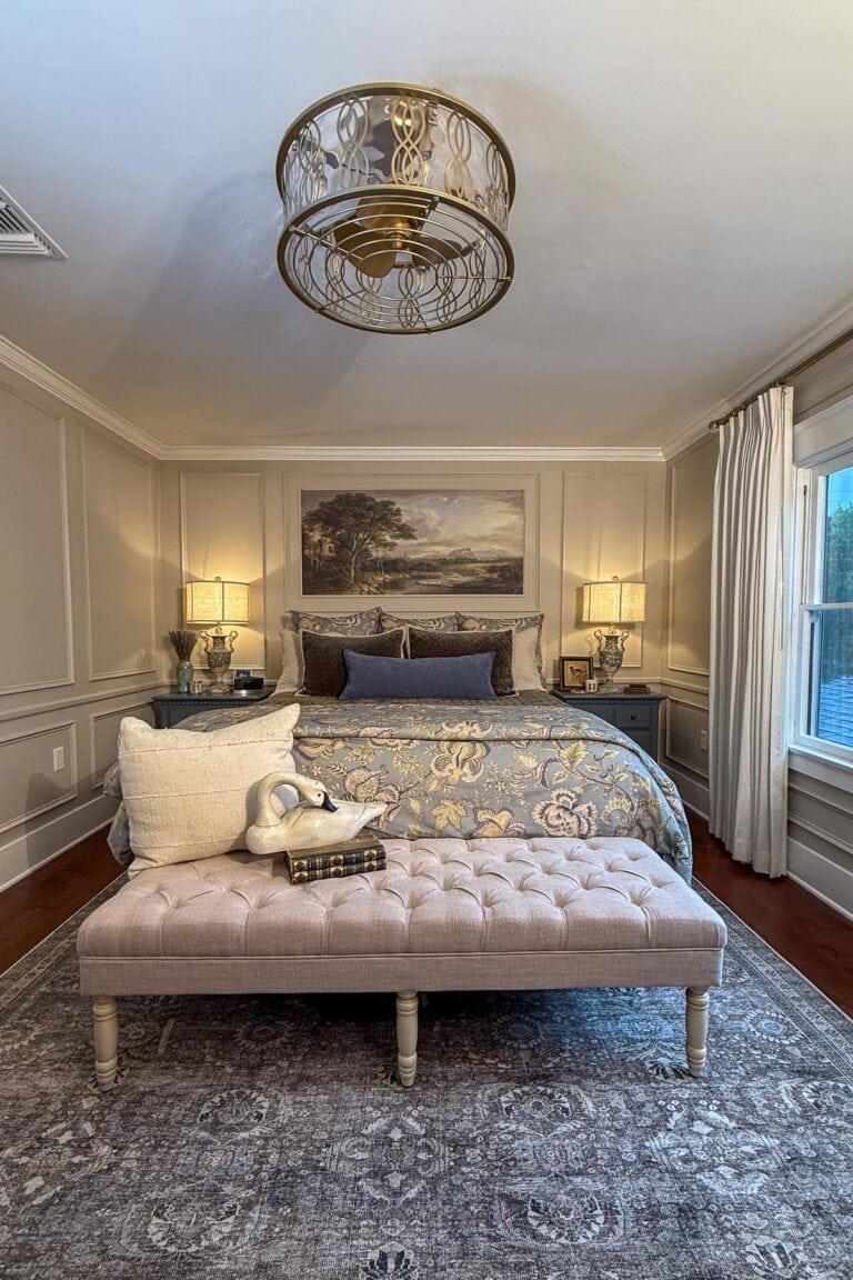 Elegant bedroom with a bed featuring floral bedding, two bedside tables with lamps, a decorative bench at the foot of the bed, and a large landscape painting above the headboard. A large window lets in natural light.