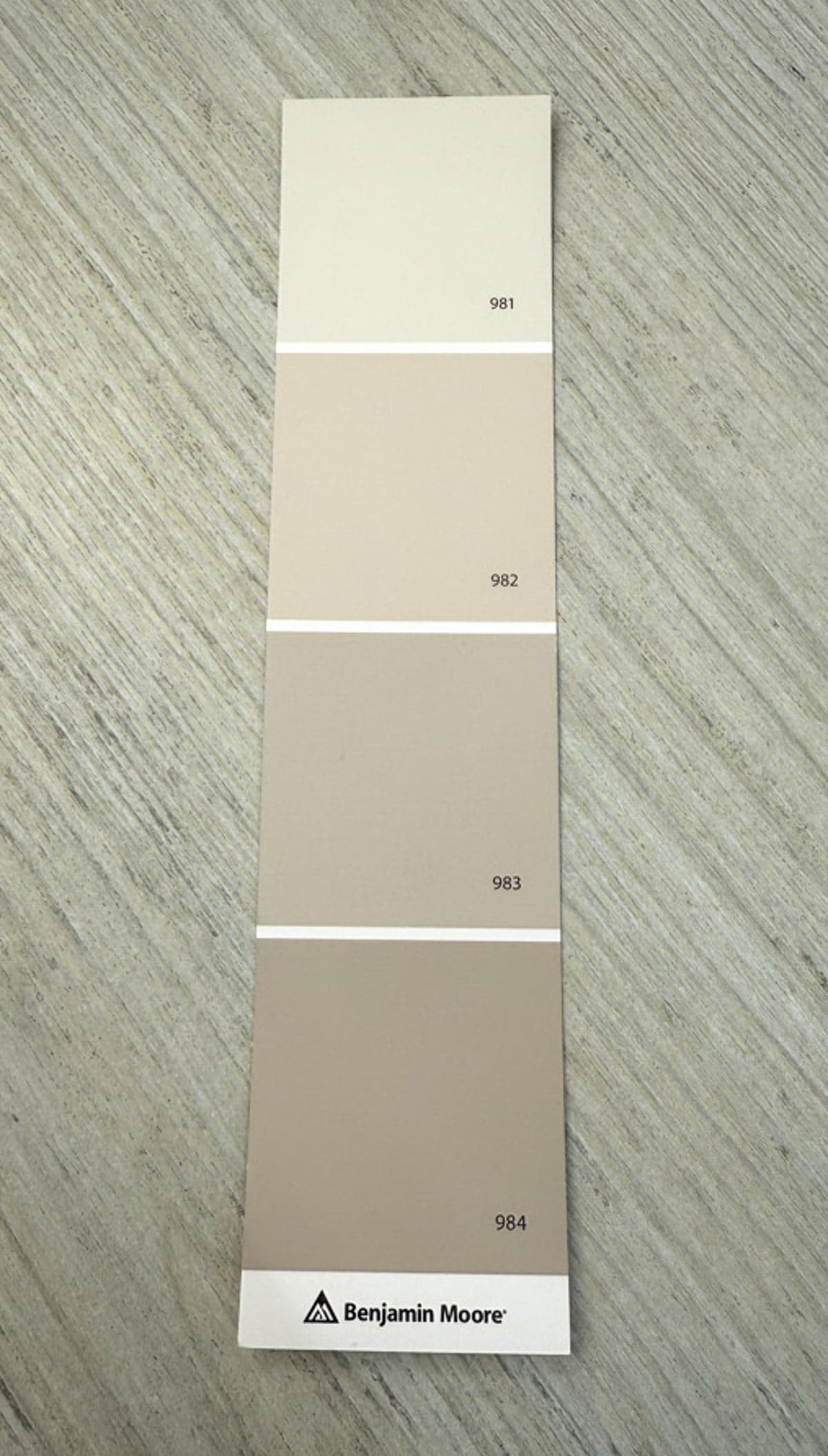 A Benjamin Moore paint swatch with four shades of beige labeled 981, 982, 983, and 984, laid on a light wood surface.