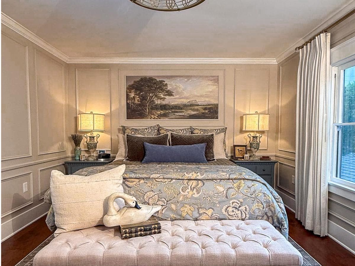 Elegant bedroom with a tufted bench at the foot of a neatly made bed featuring patterned bedding. Benjamin Moore Smokey Taupe Paint enhances the walls, while a swan-shaped towel with a book rests on the bench and artwork hangs above the bed.