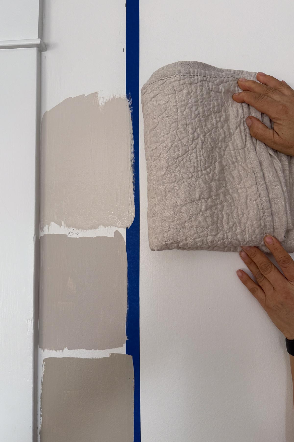 Three paint swatches in shades of beige are tested on a white wall, separated by blue tape. On the right, a person holds a folded, textured light gray fabric against the wall. Top color is Benjamin Moore Smokey Taupe reduced at 25%
