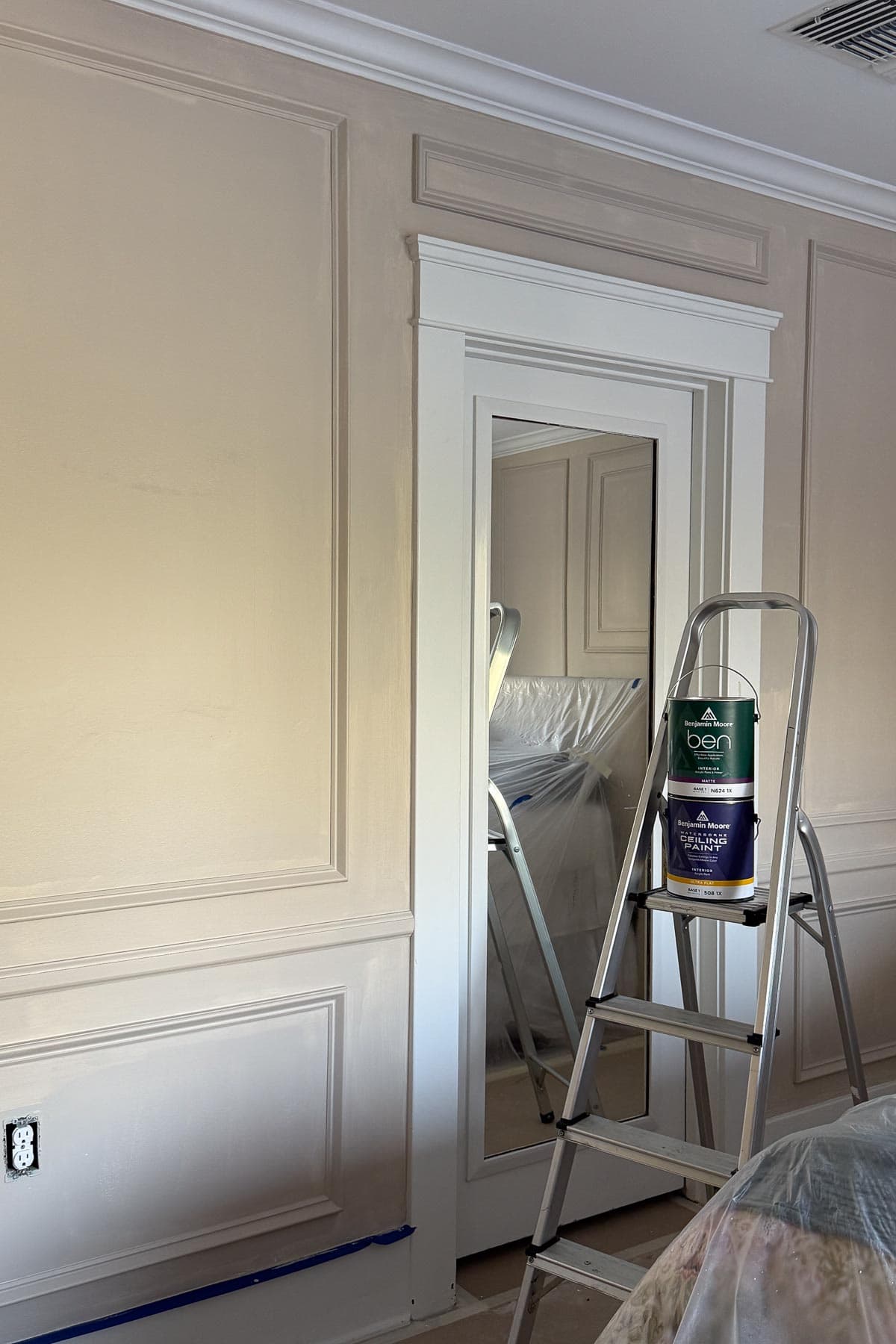 A room with beige paneled walls, a white framed door with a mirror, and a stepladder holding two paint cans. Furniture is covered with a white sheet, and painting supplies are visible on the floor.