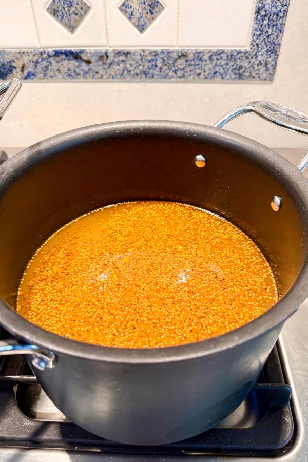 A black pot on a stove contains simmering orange-brown liquid with small visible grains, possibly quinoa, cooking. The background shows a tiled kitchen wall with blue and white patterns.