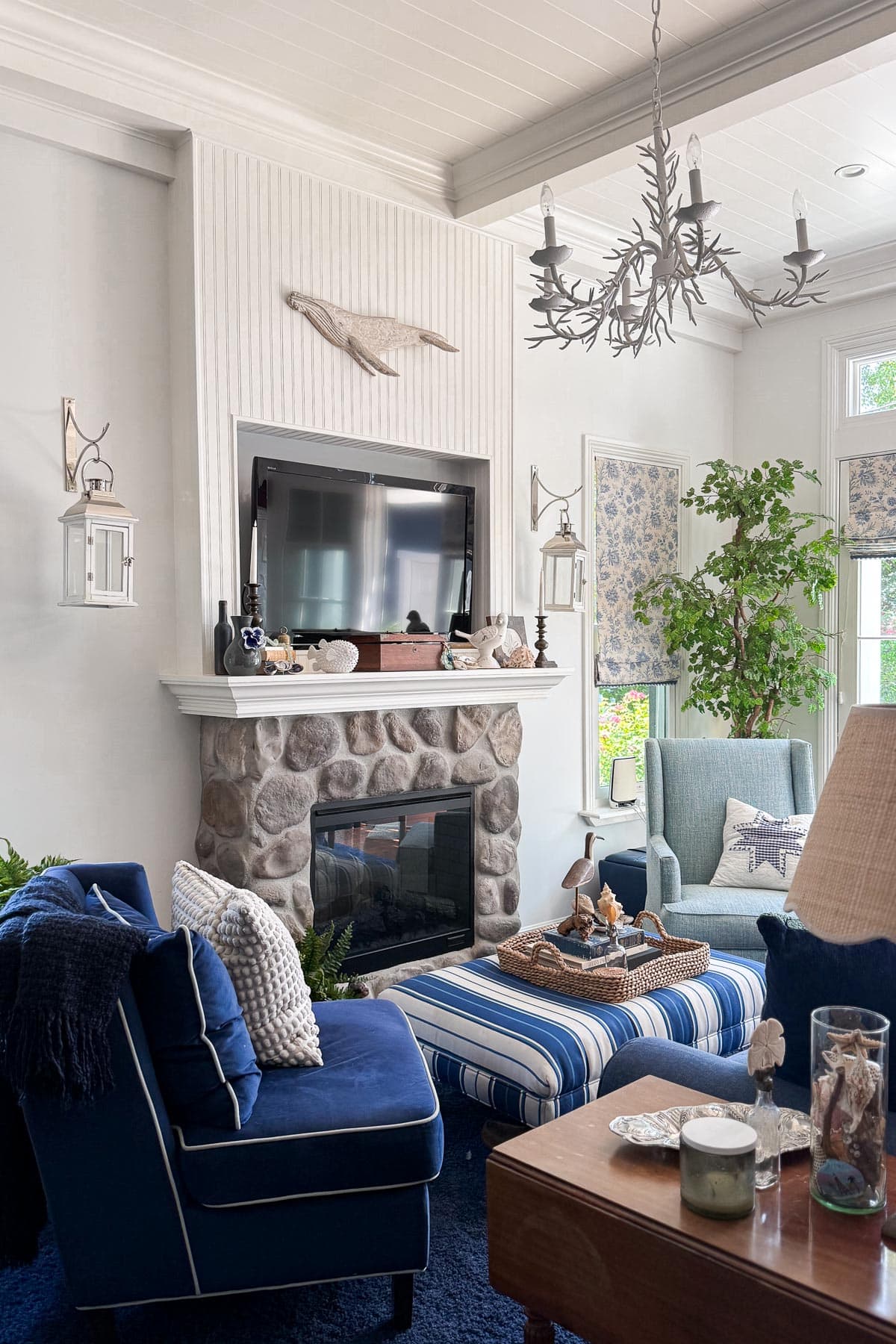 Coastal Cottage Table Decor-Cozy living room with blue velvet chairs, a stone fireplace, a TV above the mantel, nautical decor, striped ottoman, potted plants, and large windows with floral shades letting in natural light.