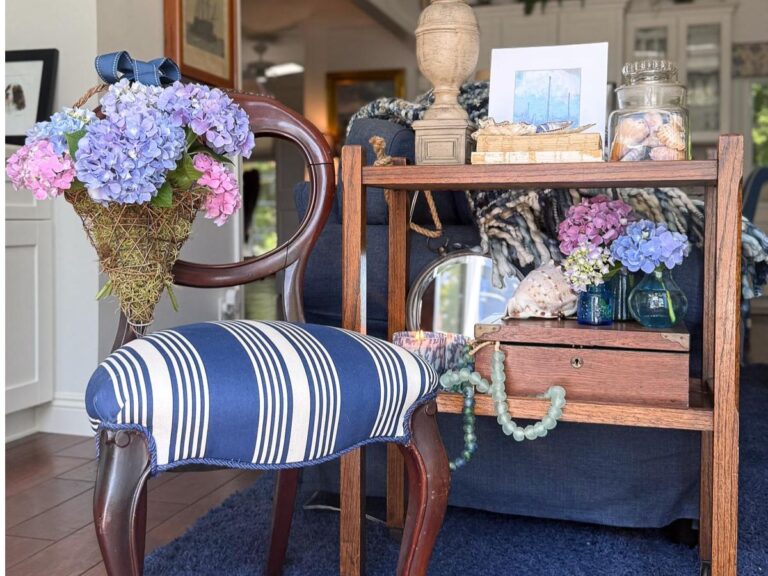 A striped blue and white chair with a bouquet of hydrangeas tied to its back stands beside a wooden shelf displaying coastal cottage table decor, seashells, a photo, books, and flowers in a cozy, well-lit living room.
