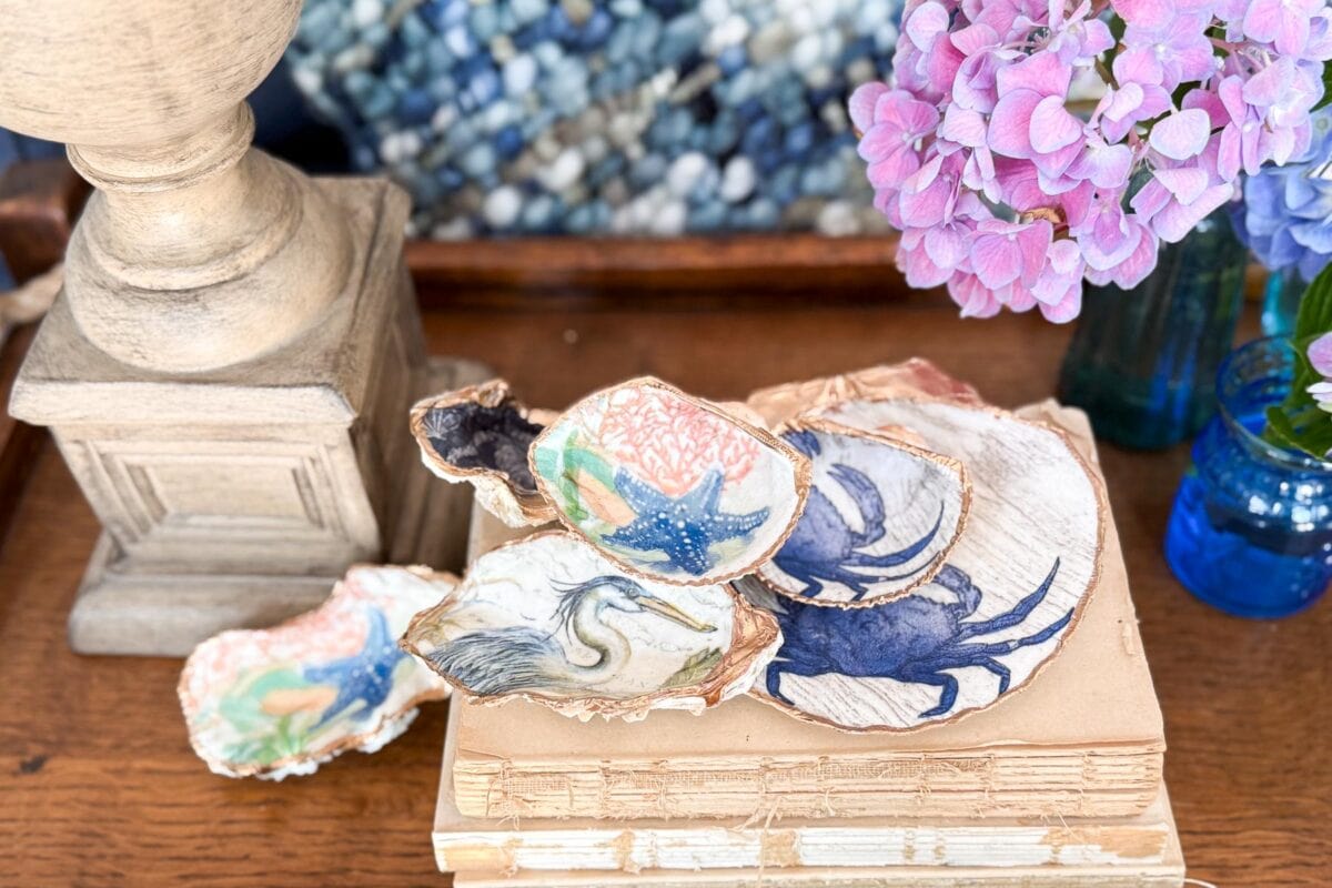 Coastal Cottage Table Decor-Painted seashells with sea creatures and birds rest on a stack of vintage books, beside a wooden lamp base and a vase of pink hydrangeas on a wooden surface.