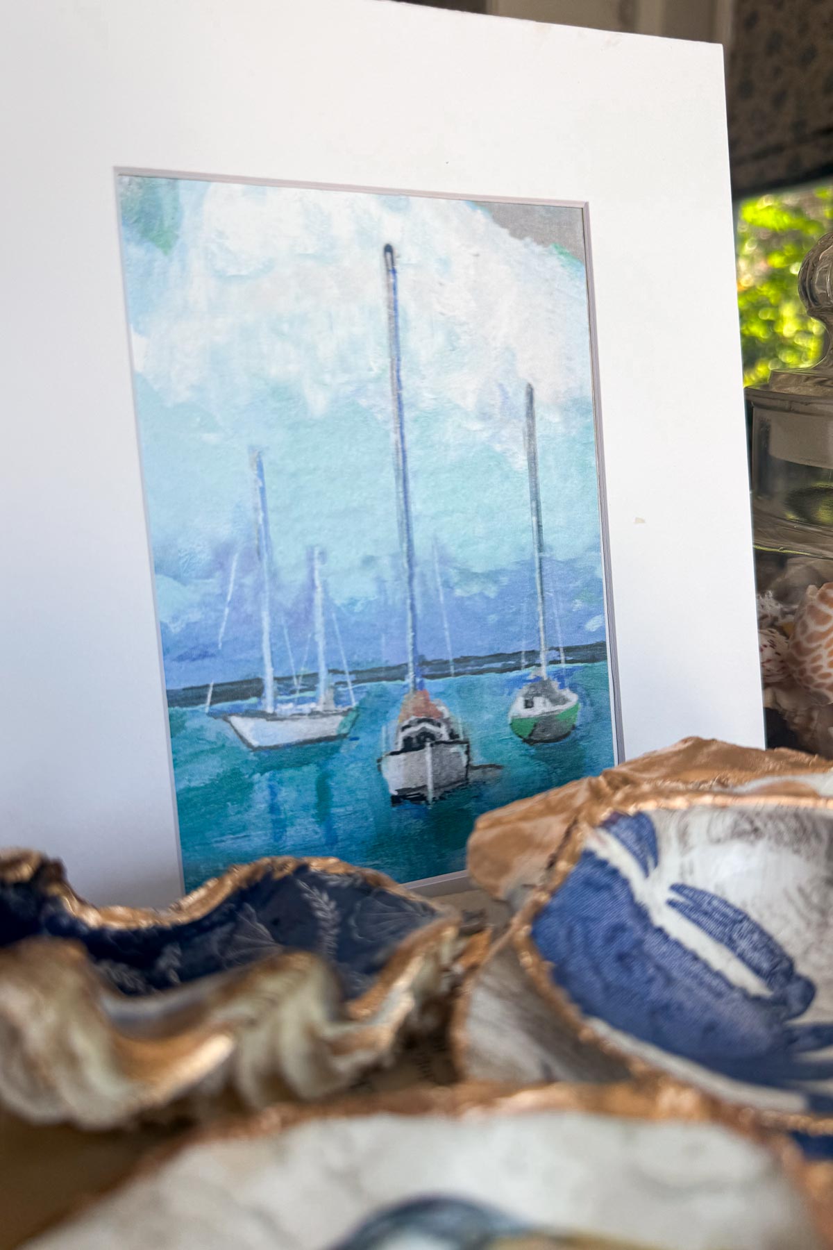 Coastal Cottage Table Decor-A framed painting of three sailboats floating on calm blue water is displayed upright, surrounded by decorative ceramic dishes with blue and gold accents. Natural light filters in from the background.