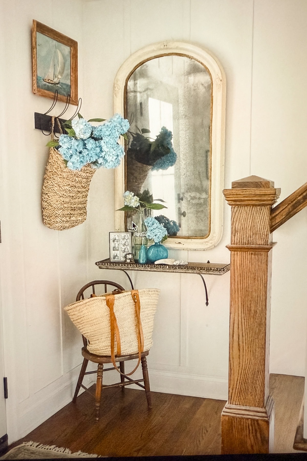 A small entryway corner with a wooden chair holding a woven tote bag, a wall shelf with blue vases, framed photos and flowers, coastal cottage table decor touches, a large vintage mirror, and a hanging basket of blue hydrangeas.