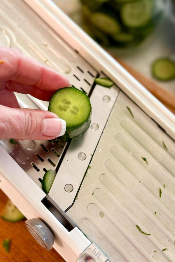 A hand holding a cucumber is slicing it on a metal mandoline slicer, with thin cucumber slices and small pieces visible on the slicer surface.