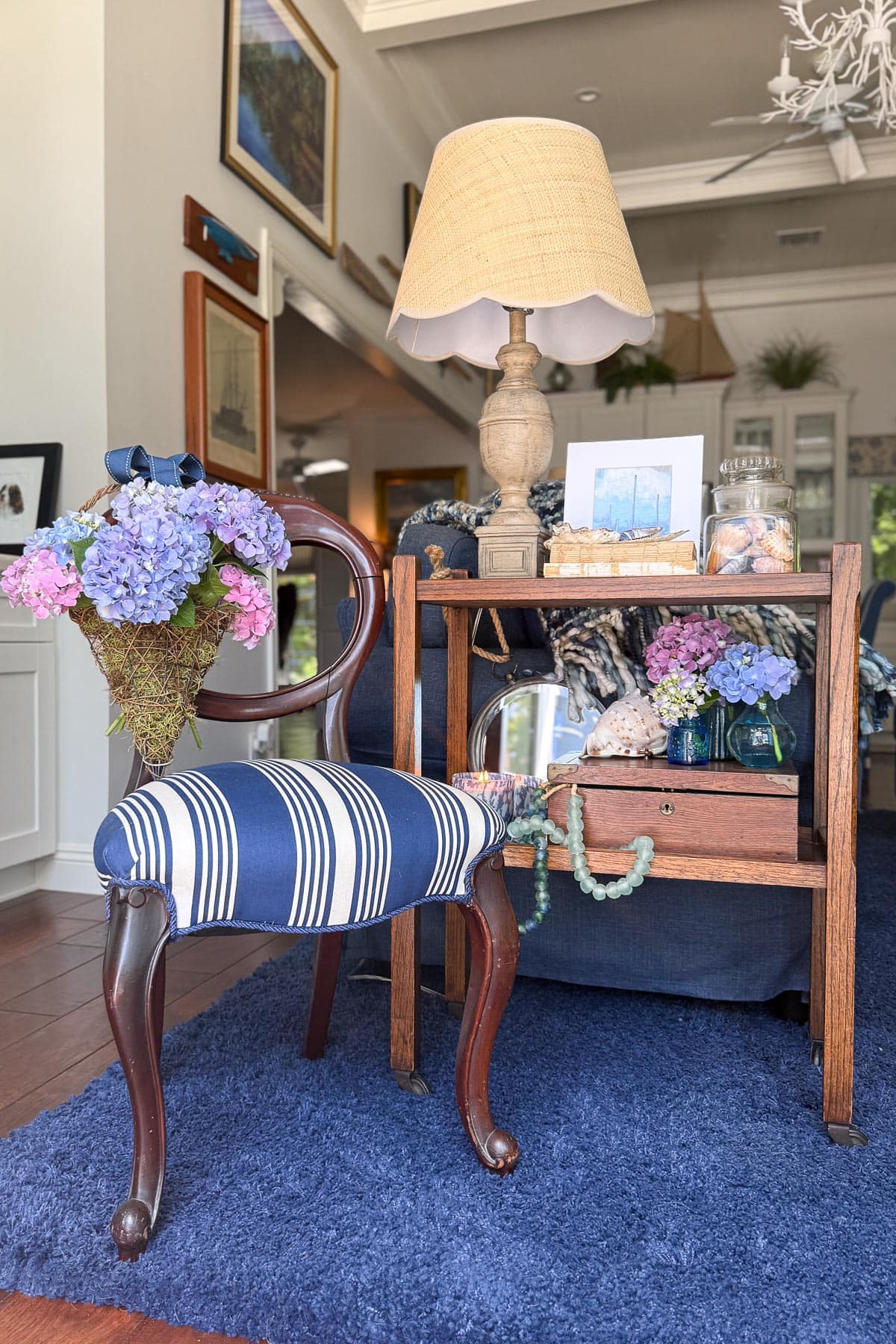 Coastal Cottage Table Decor-A cozy living room with a wooden chair featuring blue-and-white striped upholstery, a side table with a beige lamp, floral bouquet, shells, a framed photo, and nautical decor on a plush blue rug.