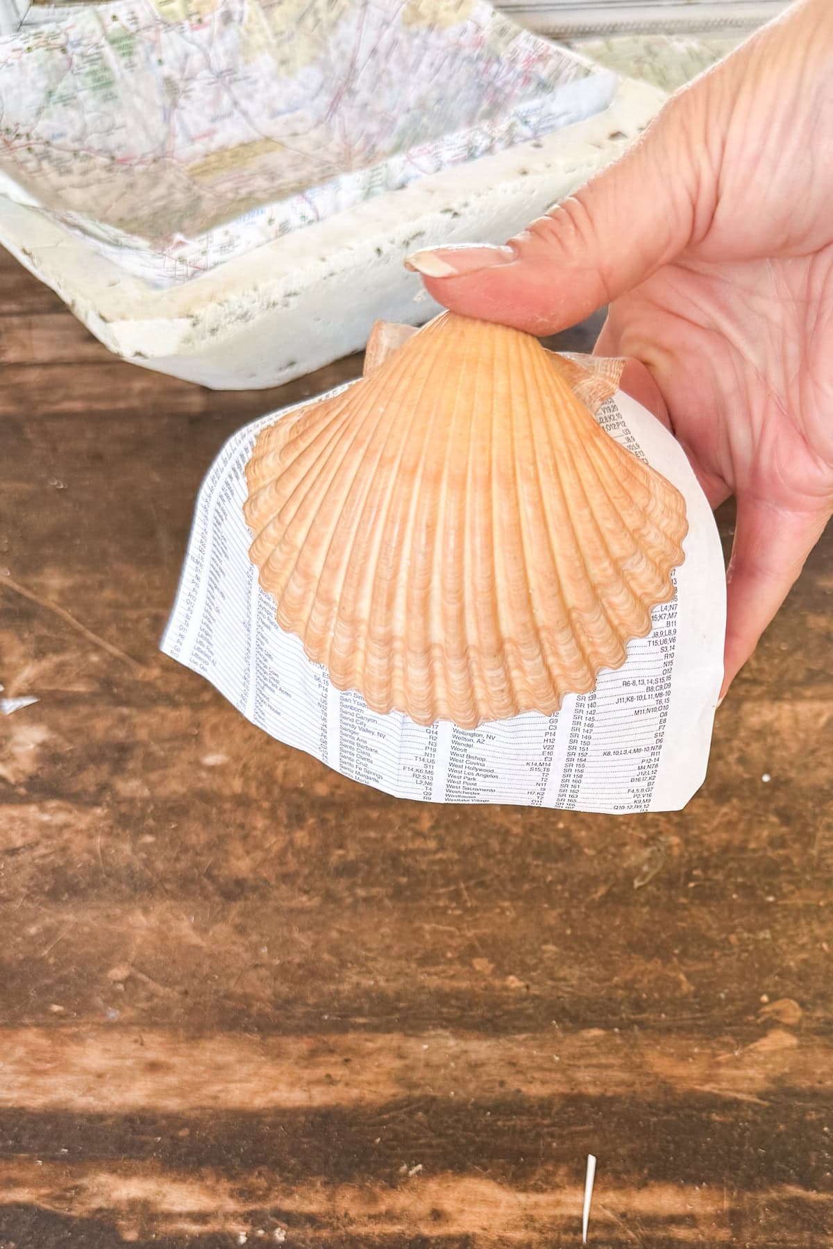 A hand holds a large seashell placed on top of a curved piece of paper with printed text, resting on a wooden surface. Craft supplies are visible in the background.