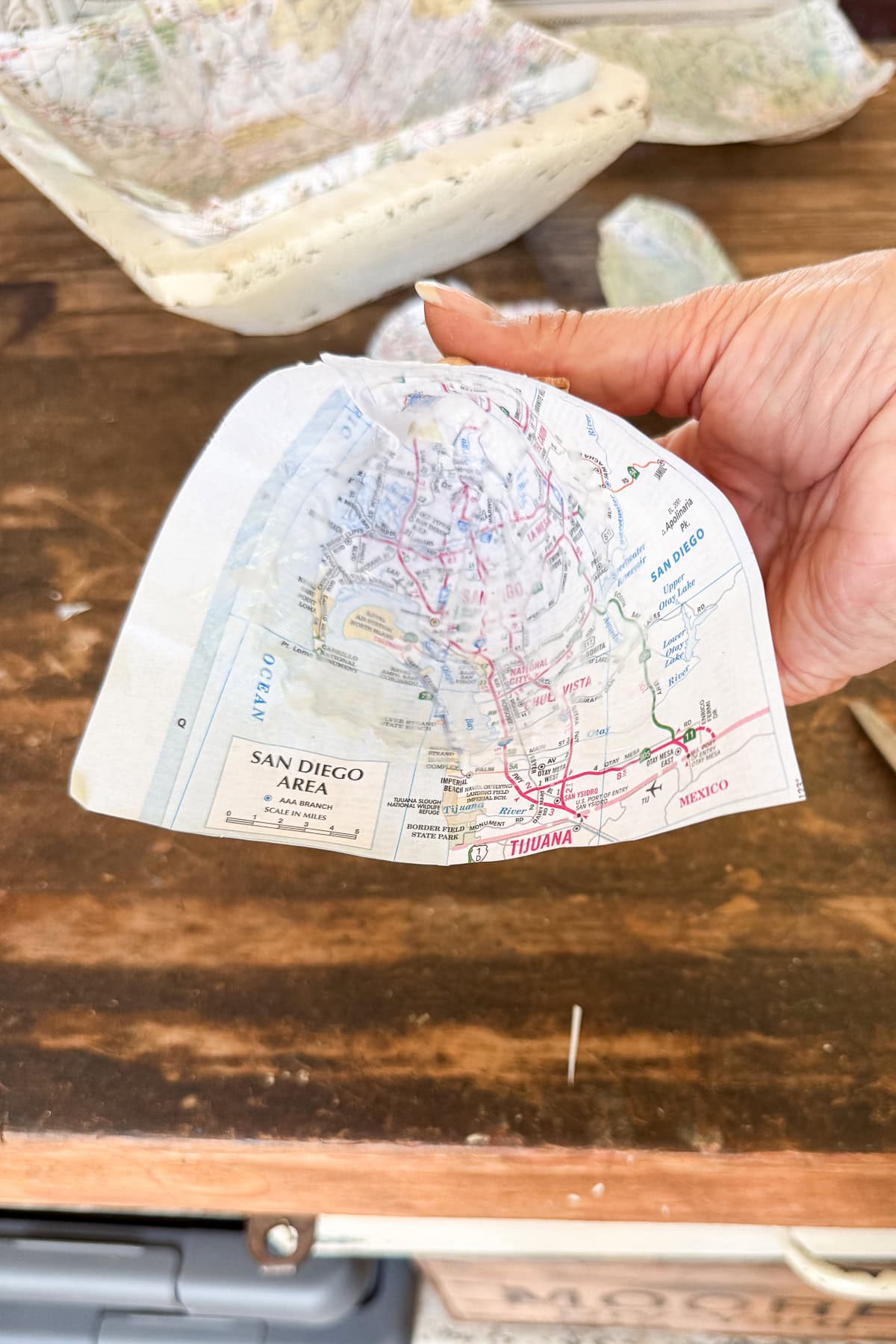 A hand holds a curved piece of paper with a map showing the San Diego area, including parts of Tijuana, Mexico, and surrounding roads, over a wooden table with other map pieces in the background.