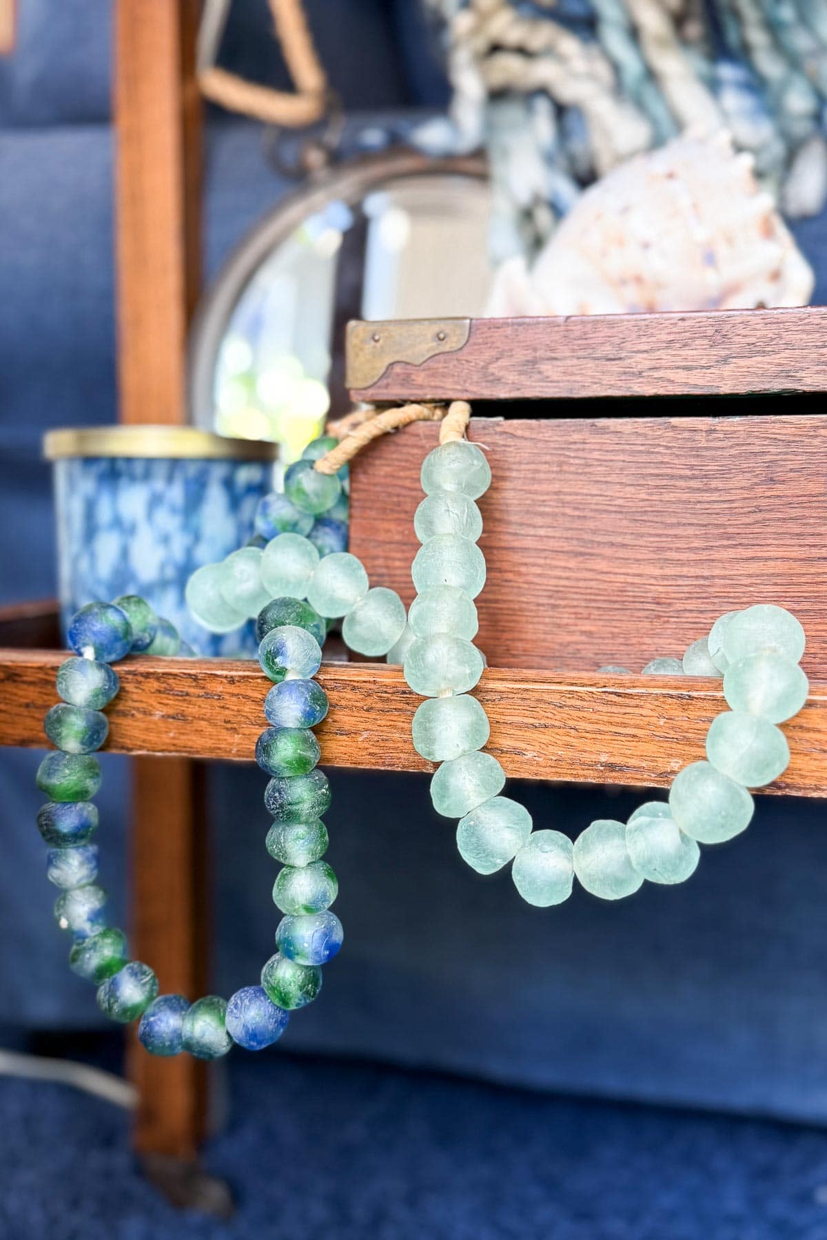 Two beaded glass necklaces, one green and one blue-green, hang from the edge of a wooden box. In the background, a blue container, a mirror, and a seashell are partially visible.