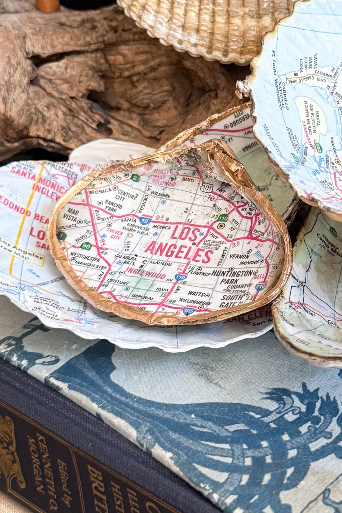 A decorative decoupage shell dish features a map of Los Angeles with gold-painted edges, resting on a book and surrounded by other Decoupage Shells with Maps and natural elements.
