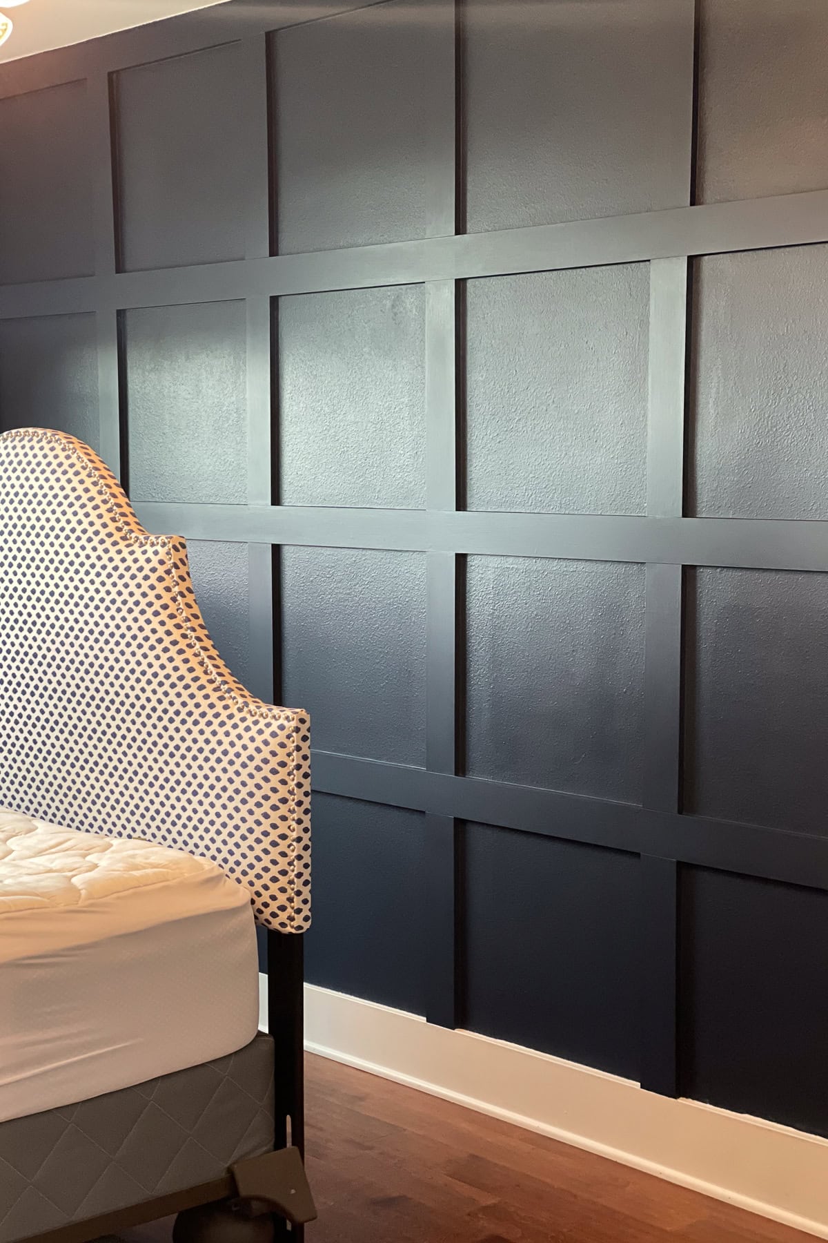 A navy blue accent wall with a grid panel design beside a bed with a white and blue patterned upholstered headboard and a white mattress on a wooden floor.