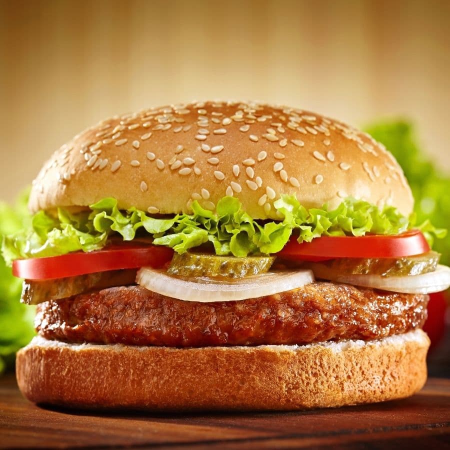 A close-up of a hamburger with a sesame seed bun, lettuce, tomato slices, Mrs. Wages Bread and Butter Pickles, onions, and a cooked beef patty on a wooden surface, with a blurred background.