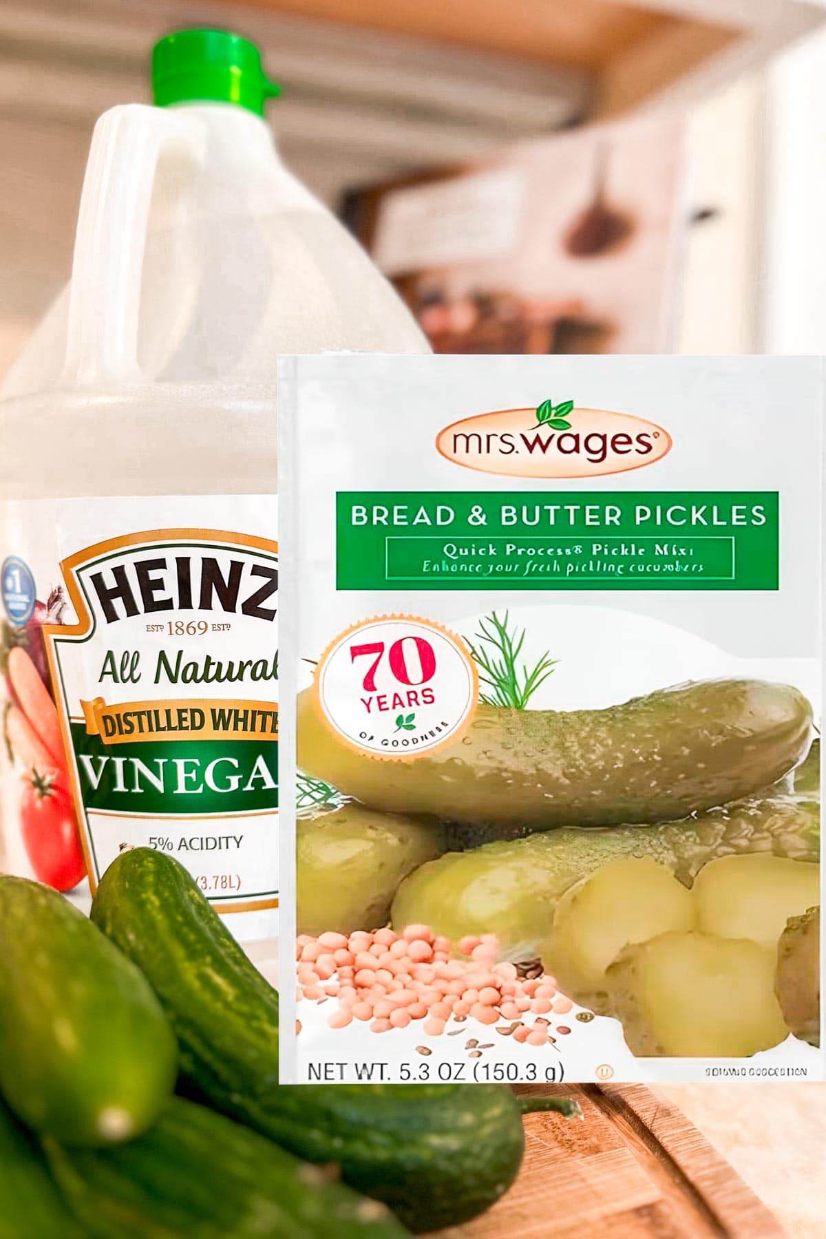 A bottle of Heinz distilled white vinegar, a packet of Mrs. Wages bread & butter pickles mix, and fresh cucumbers are arranged on a kitchen counter.
