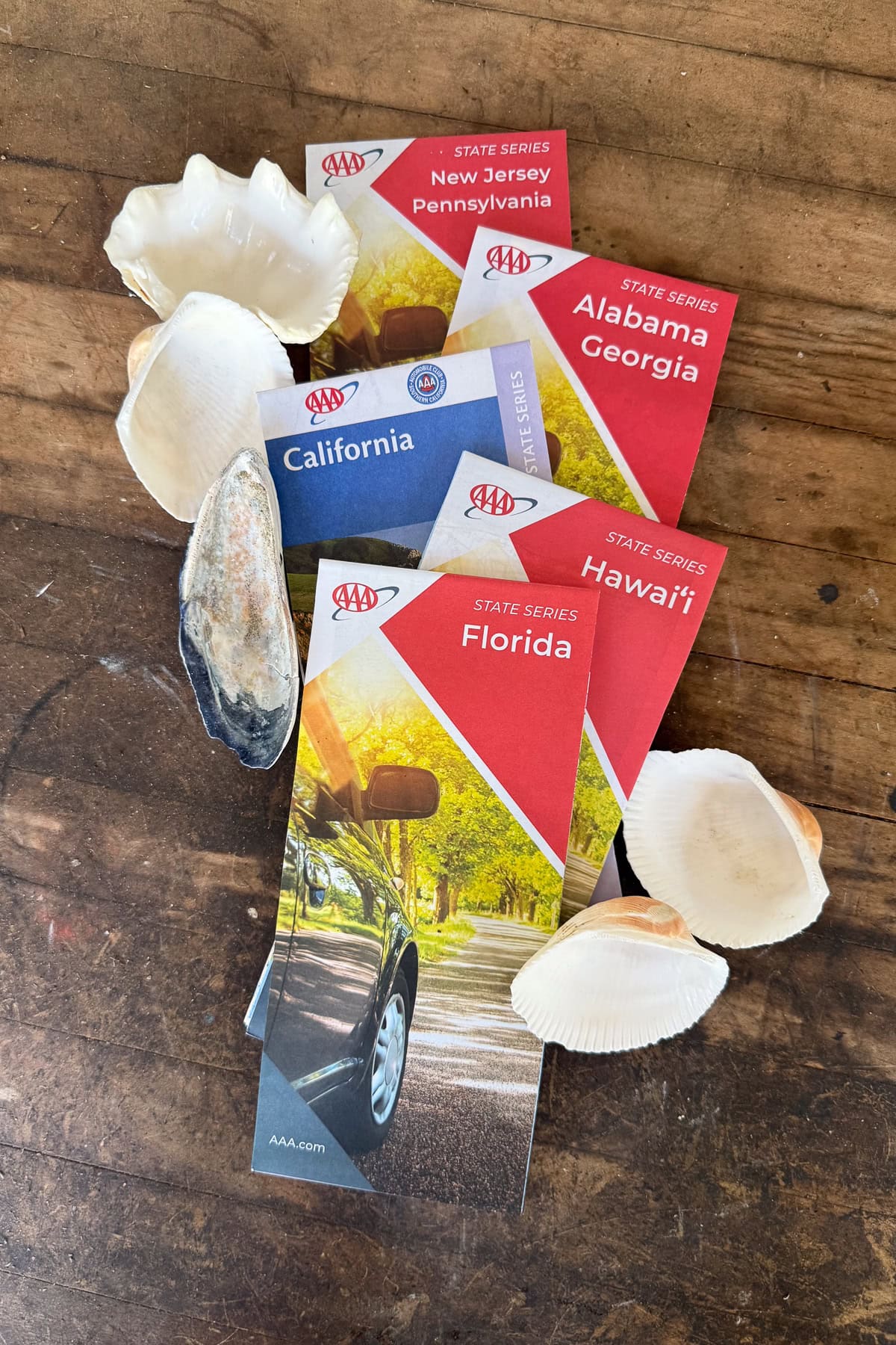 Five AAA state travel guides—New Jersey/Pennsylvania, Alabama/Georgia, California, Florida, and Hawaii—are spread on a wooden surface, surrounded by various seashells.