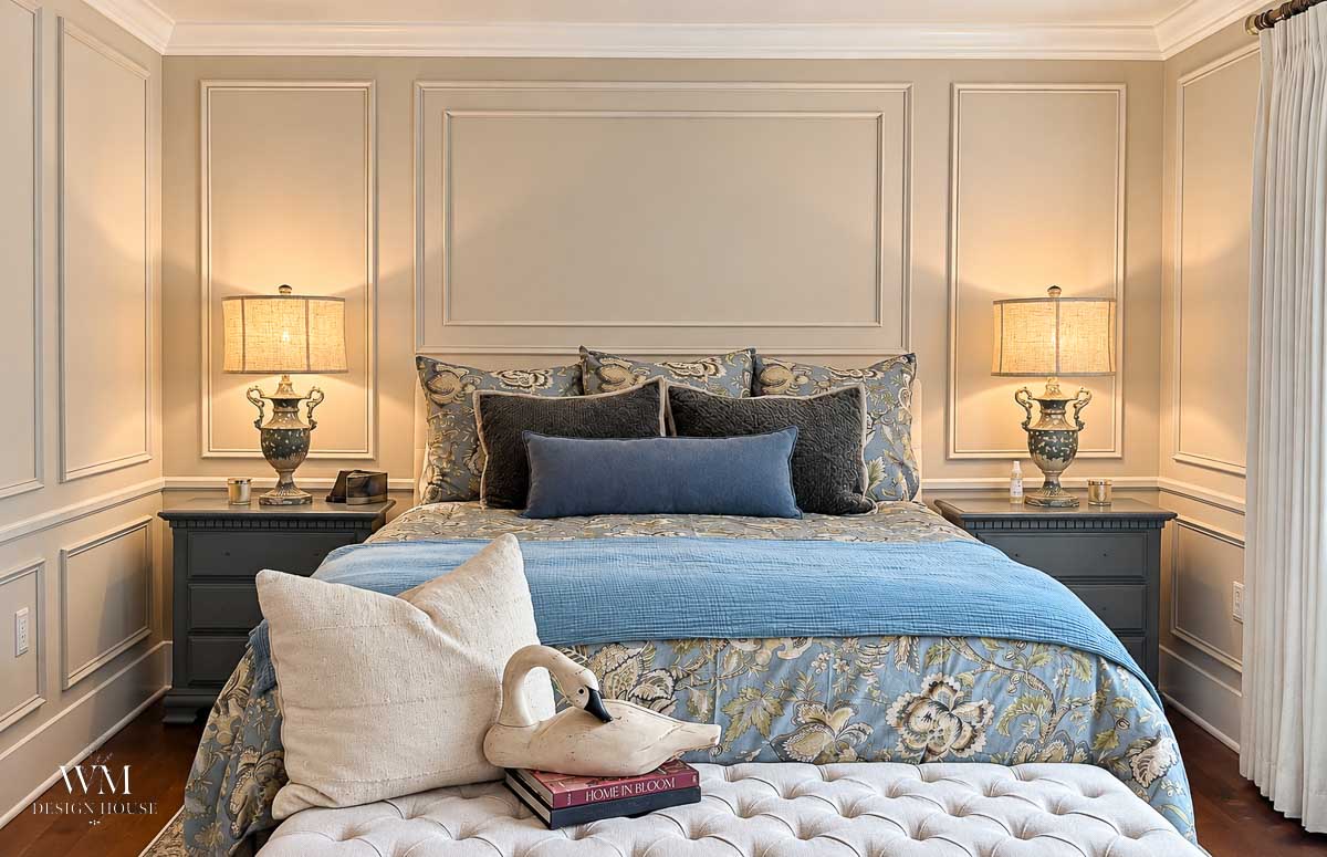 Elegant bedroom with a patterned bed, blue and beige bedding, two bedside tables with lamps, and a white bench holding throw pillows, books, and a folded towel shaped like a swan. The walls are painted in Benjamin Moore Smokey Taupe Paint, adding a sophisticated and calming backdrop to the serene space.