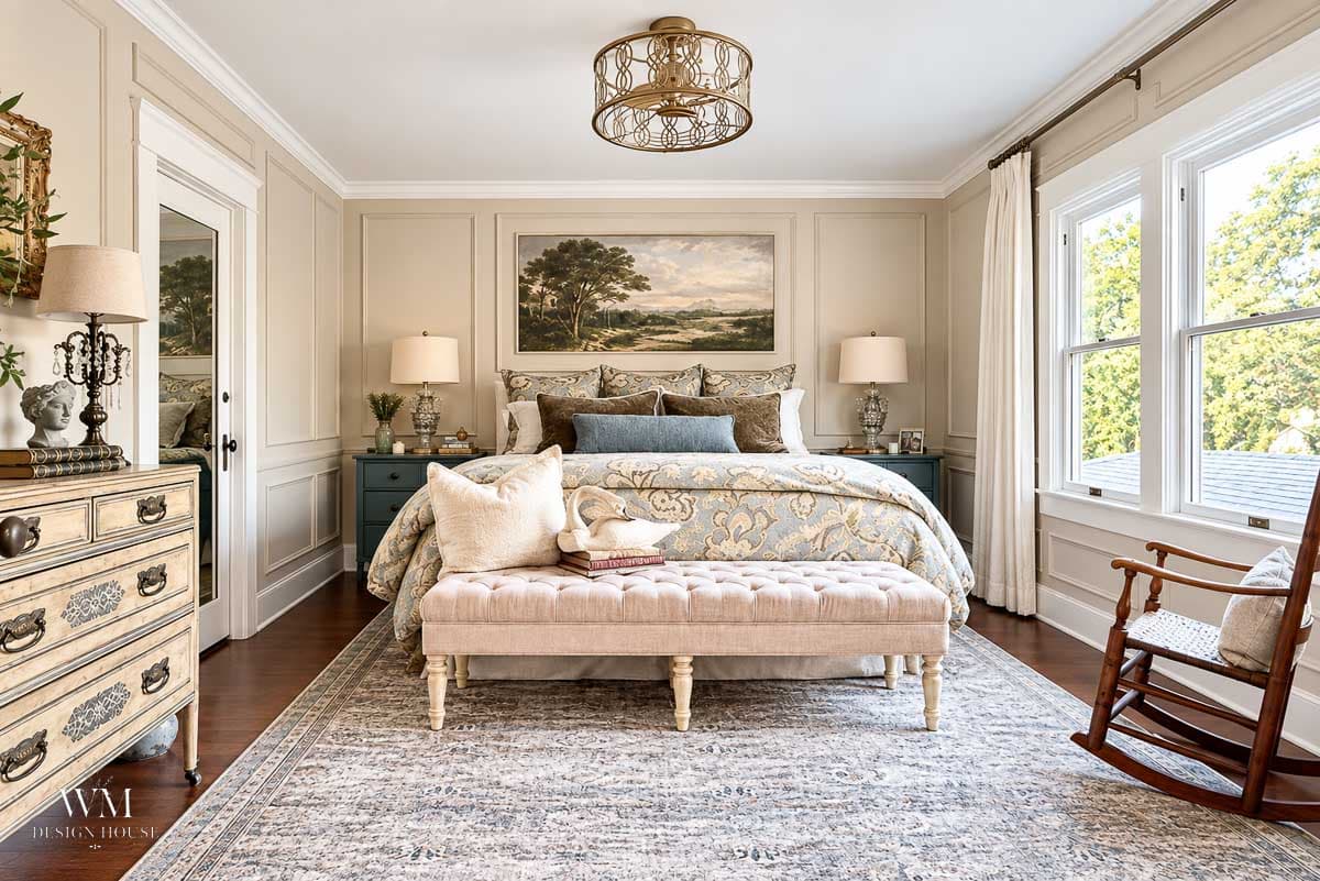 Elegant bedroom with a king bed, tufted bench, patterned rug, and two nightstands. Large windows let in natural light, beautifully highlighting the walls painted in Benjamin Moore Smokey Taupe. Neutral tones, framed art above the bed, and a wooden rocking chair complete the cozy decor.