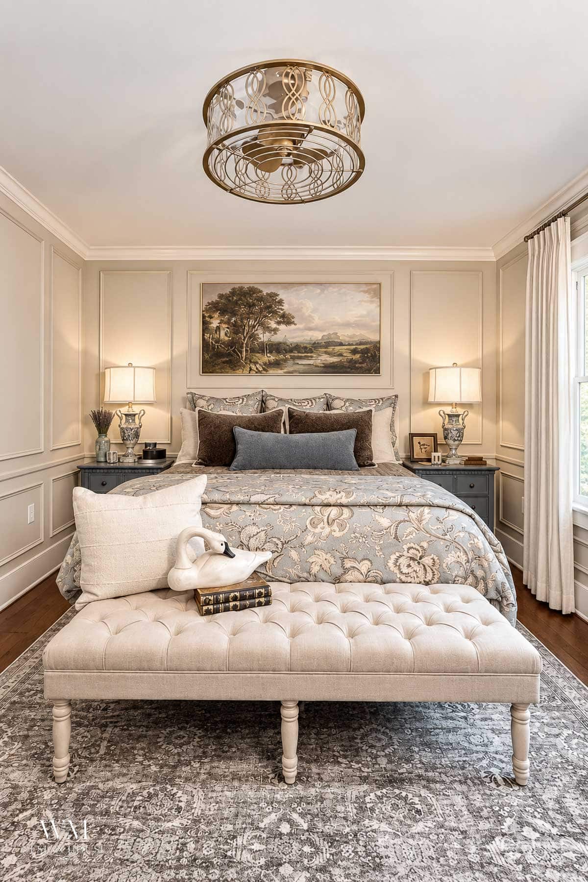 Elegant bedroom with a tufted bench at the foot of a bed dressed in floral bedding, two bedside tables with lamps, and a large decorative ceiling light fixture above. Walls painted in Benjamin Moore Smokey Taupe Paint create a soothing backdrop, while a landscape painting hangs over the bed.